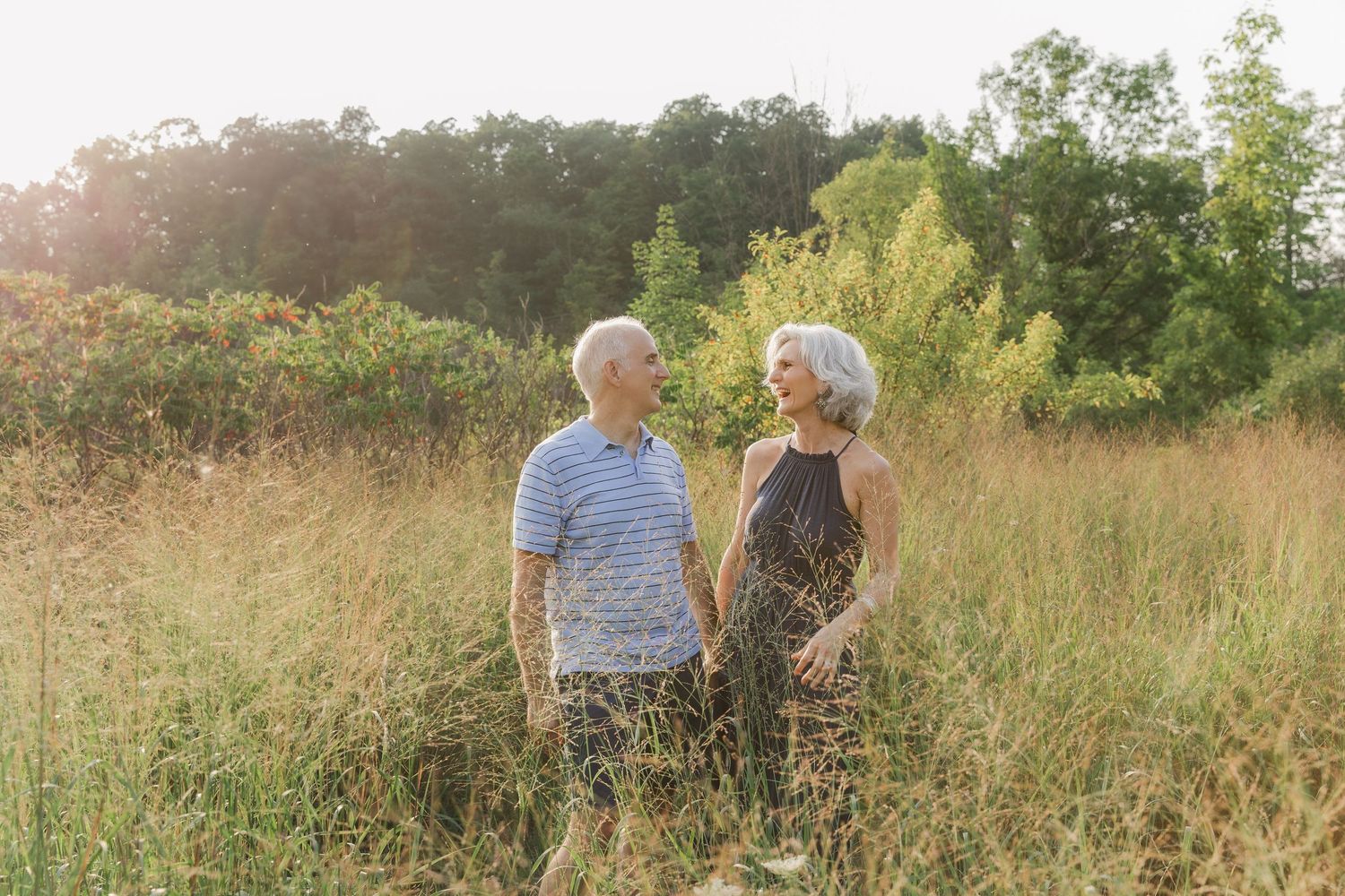 A couple walks through a golden field of tall grass during sunset while holding hands and enjoying nature together.