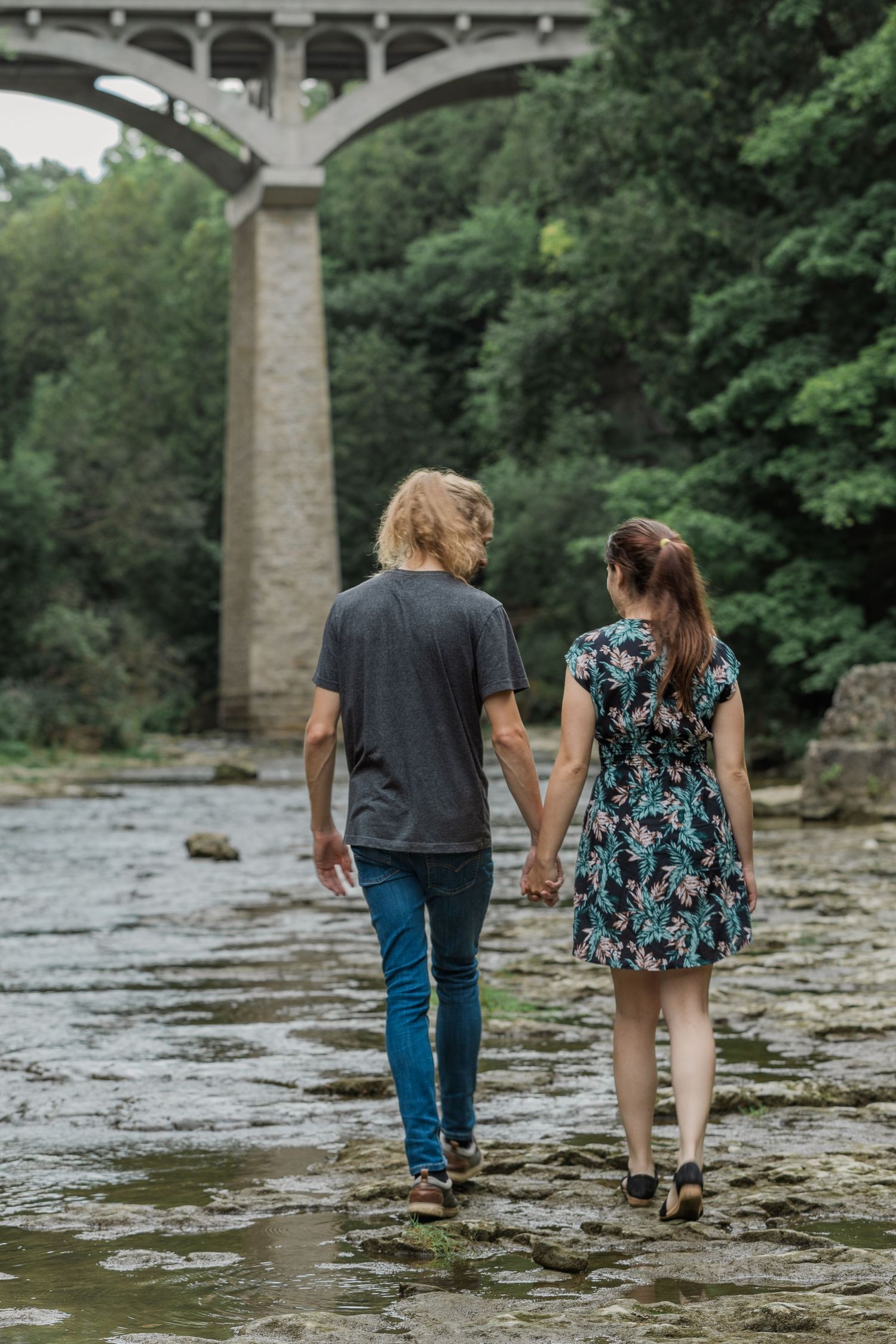 A couple walks hand in hand along a rocky riverbed with stone bridge pillars visible in the background.