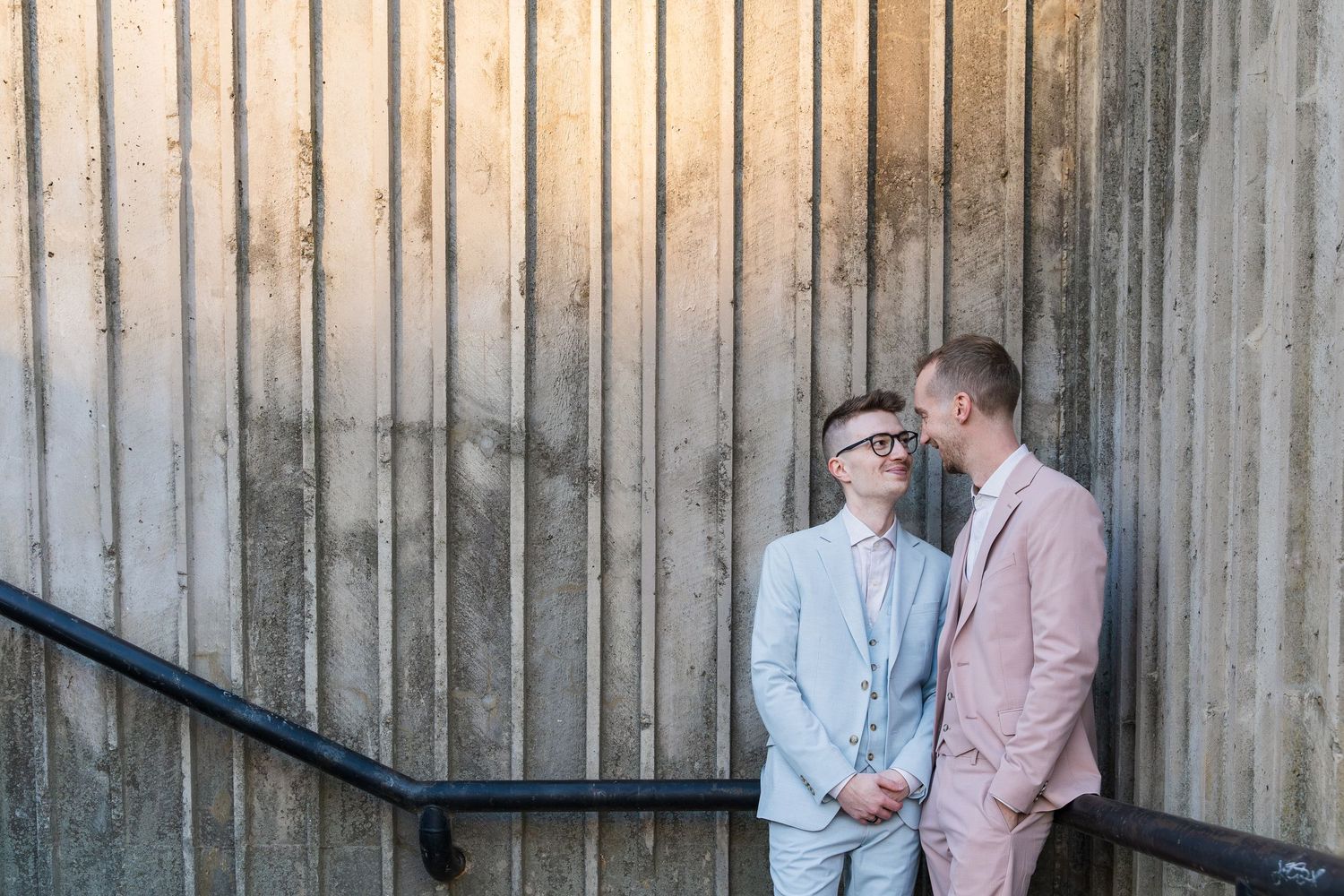 Groom and groom looking lovingly at each other in a stairwell wearing blue and pink suits by the river in downtown galt, Ontario.