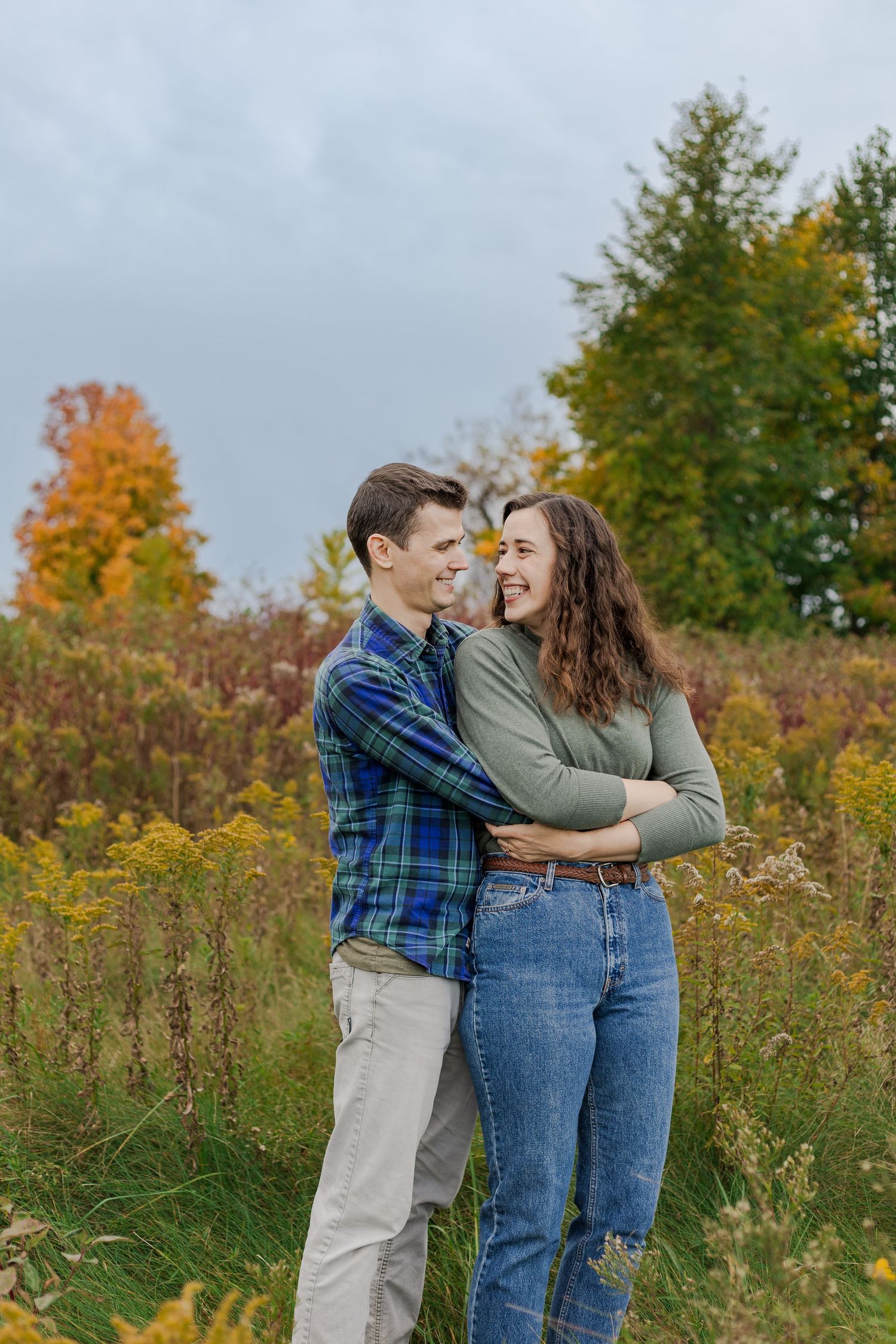 A couple embraces and laughs together in a field during autumn with colorful fall foliage in the background.