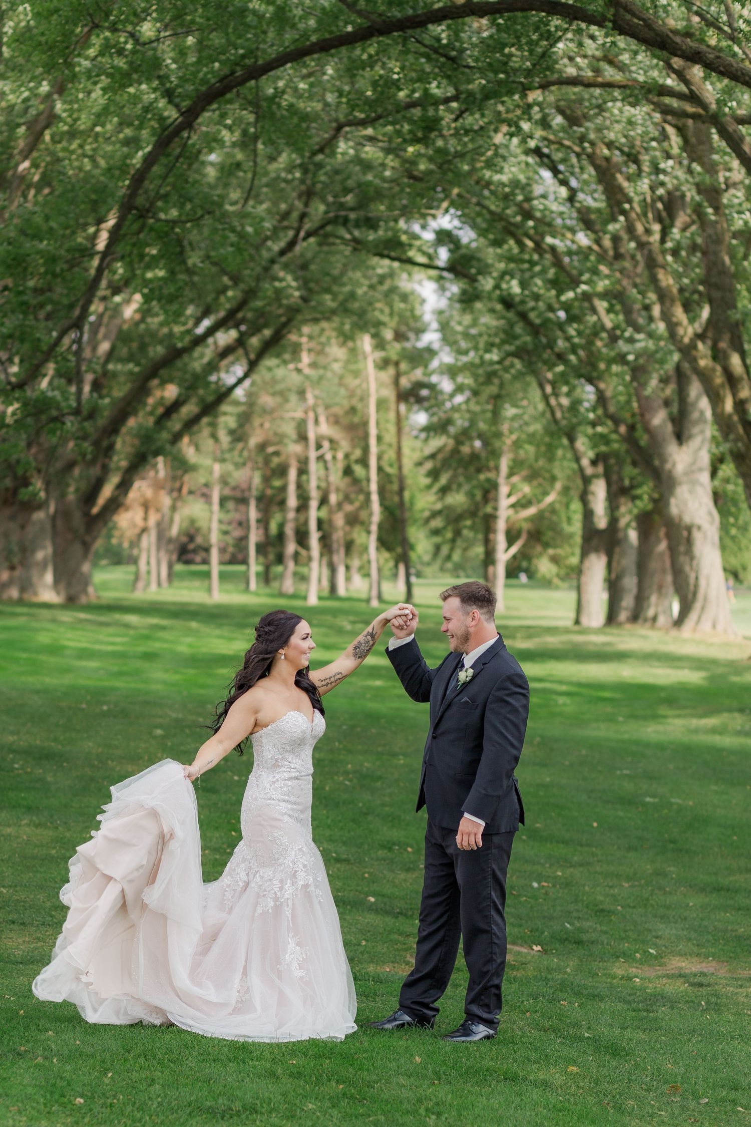 Natural wedding photo of groom twirling his bride under the canopy of rows of large trees near woodstock, ontario.