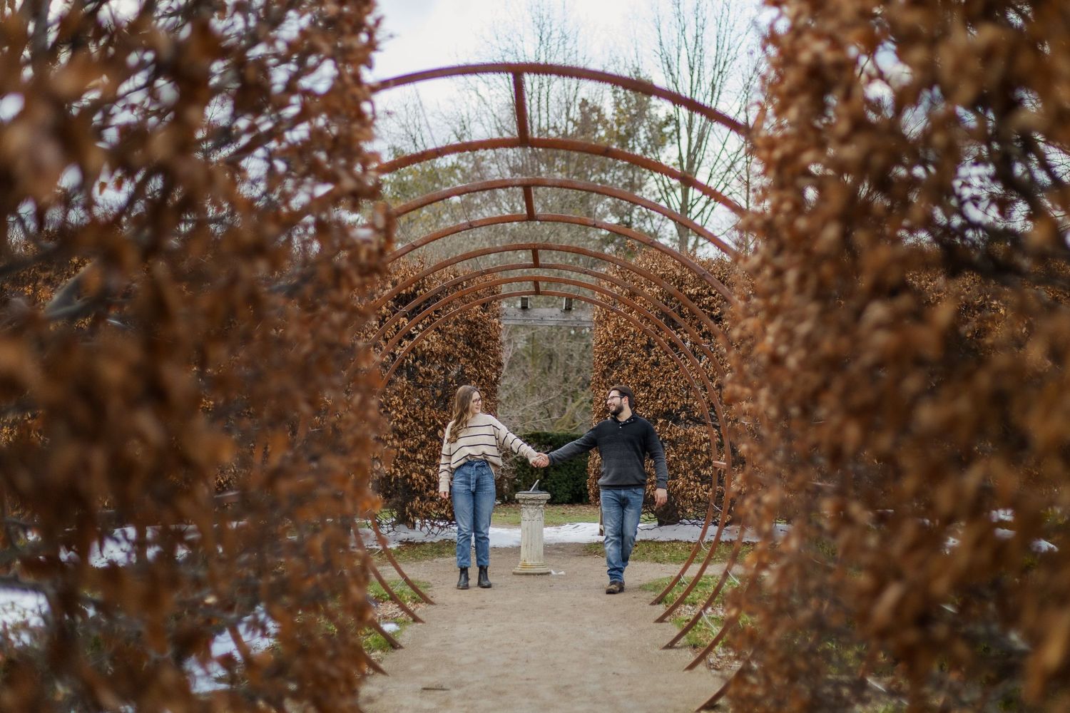 Couple holding hands walking through a circular metal structure during their winter engagement session at the guelph arboretum.