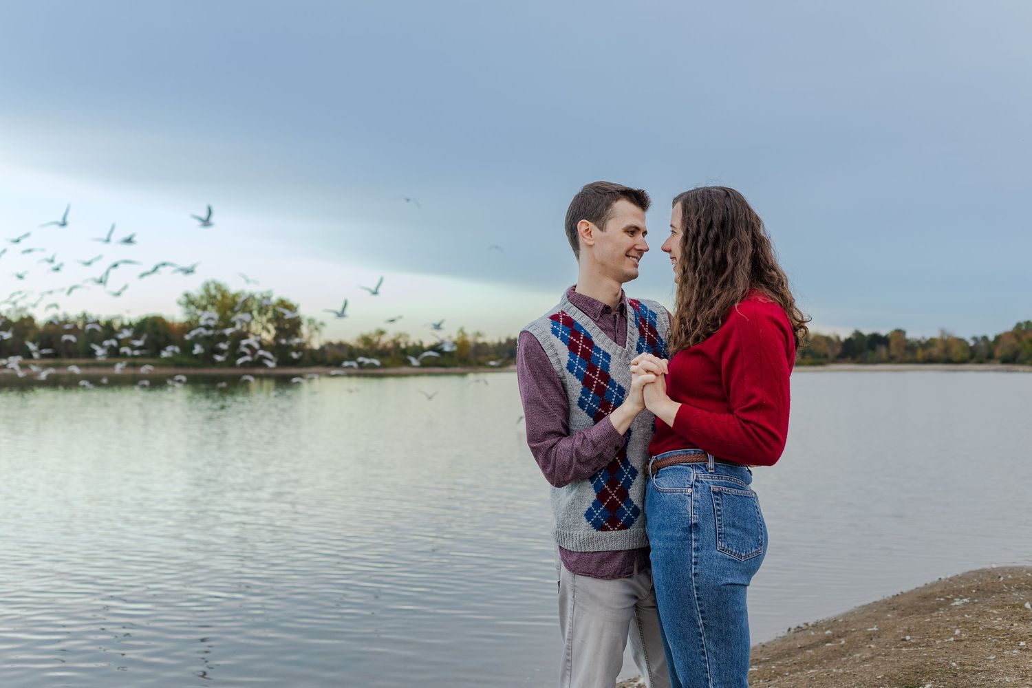 Relaxed engagement photo of couple standing close together iby the lake, birds flying in the background, at guelph lake conservation area.