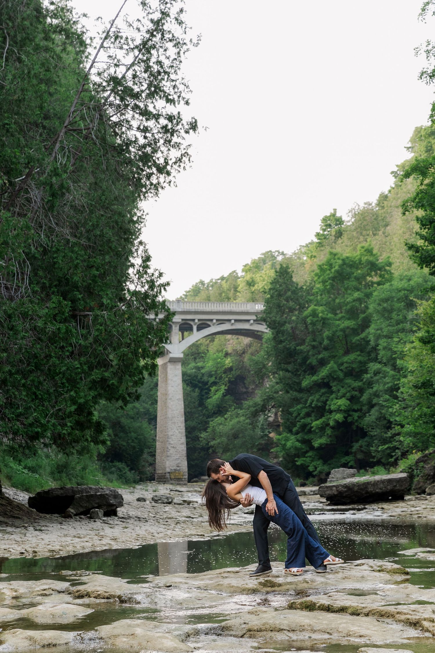 Couple performs a dip along the rocky river surrounded by tall trees with a bridge in the background at The Elora Gorge.