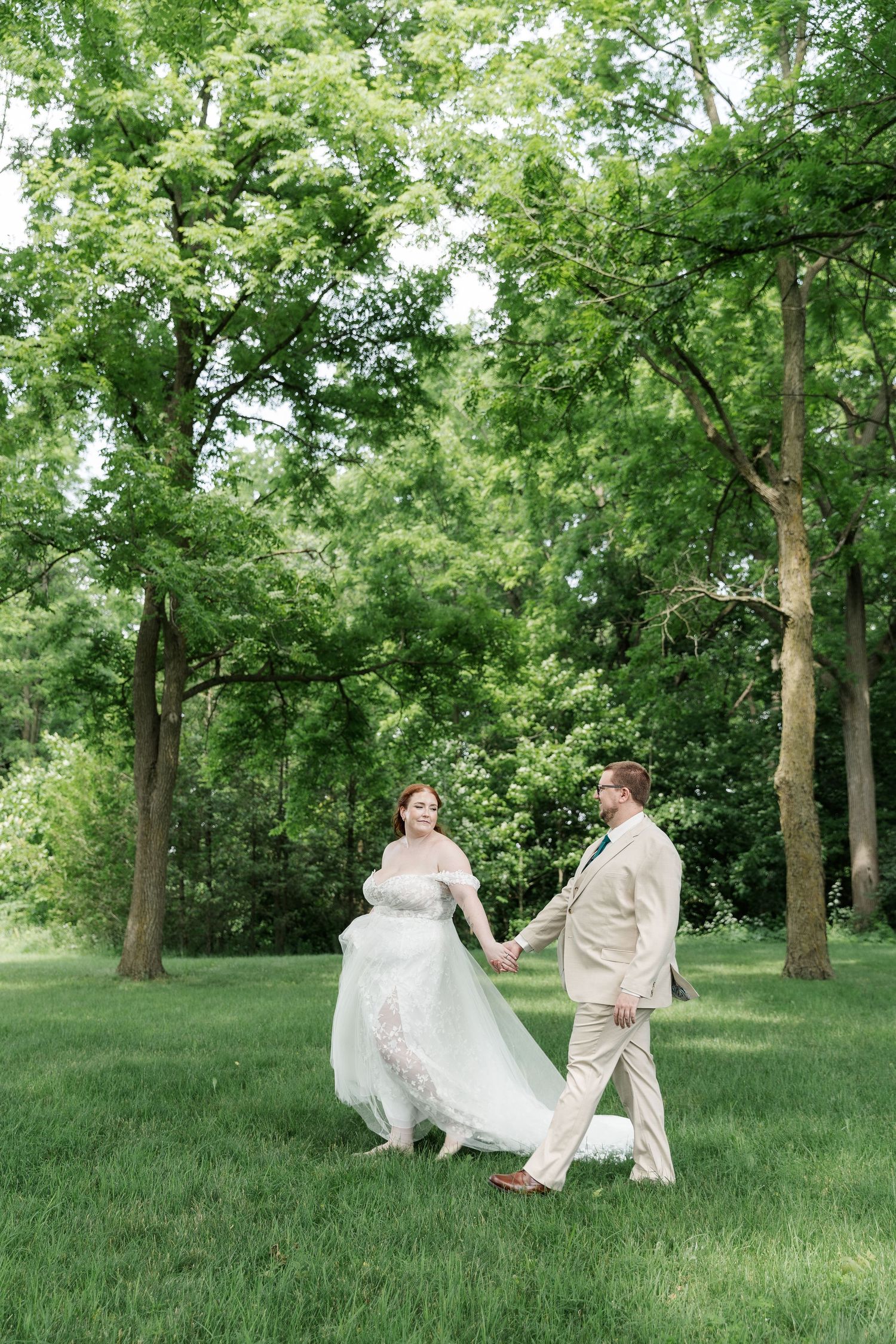 Bride and groom walking through a field surrounded by large trees during their wedding photos near woodstock, ontario.