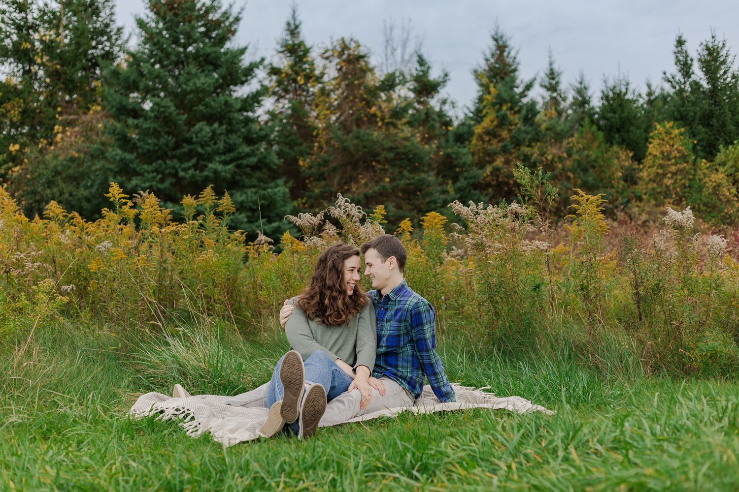 Relaxed engagement photo of couple sitting on a blanket in a field by yellow wild flowers at the guelph lake conservation area.