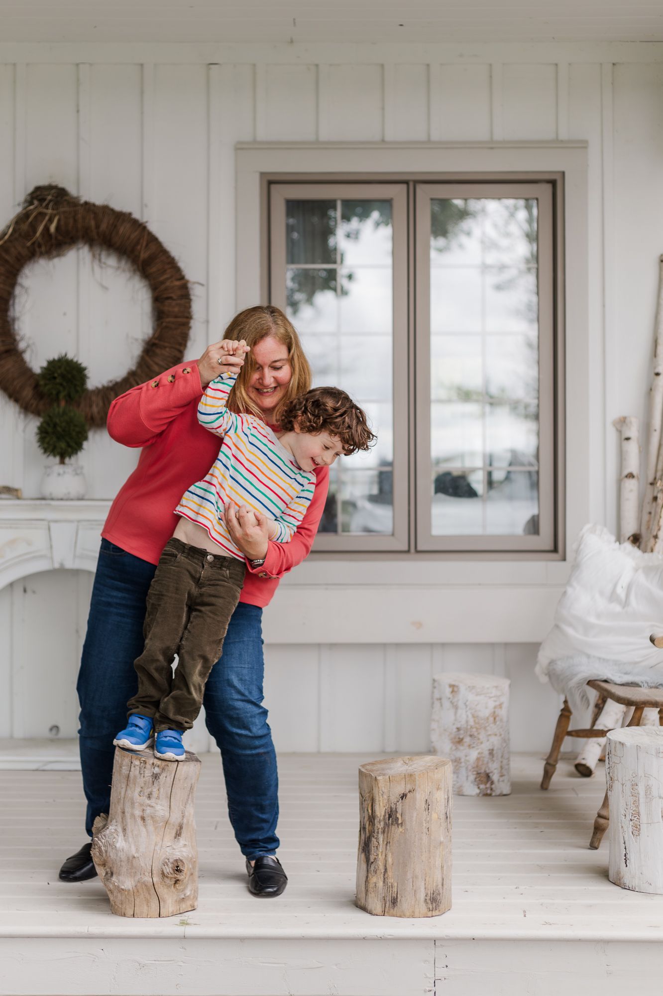A mother playing with her son on some decorative logs on the front port of the white countryside home in waterford, ontario.