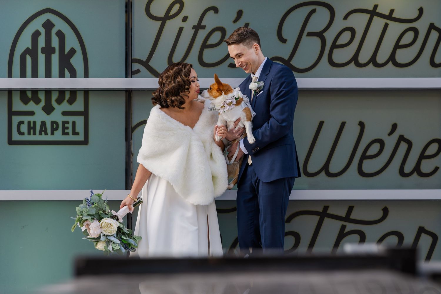 Groom holding dog while bride gives her a kiss in front of the DTK chapel in downtown Kitchener, Ontario