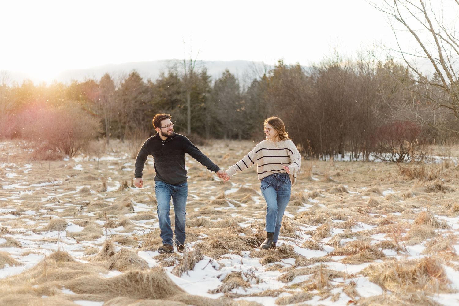 Two people walk hand in hand through a snowy winter field at sunset with bare trees in the background.
