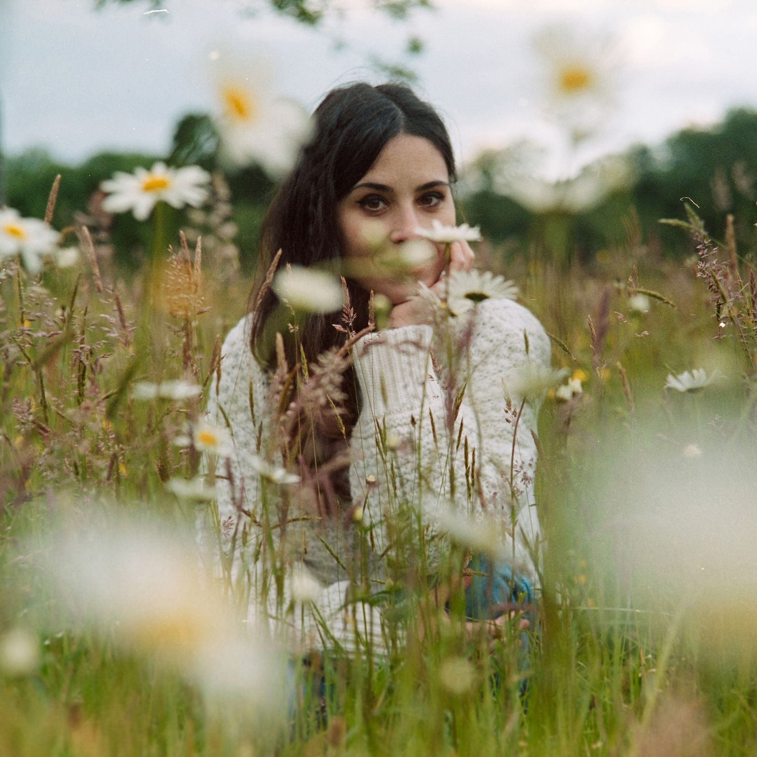 A woman in a light-colored sweater sits among blooming daisies and wildflowers in a dreamy summer meadow.