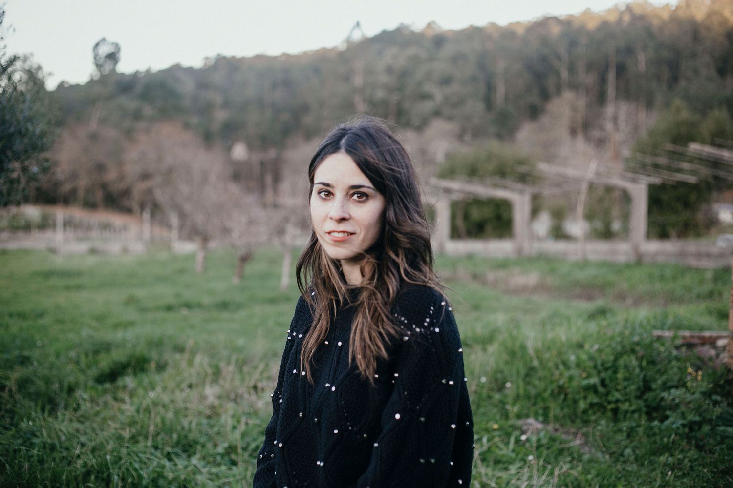 Brunette in a dark sweater standing in a grassy field with rolling hills and trees in the background.