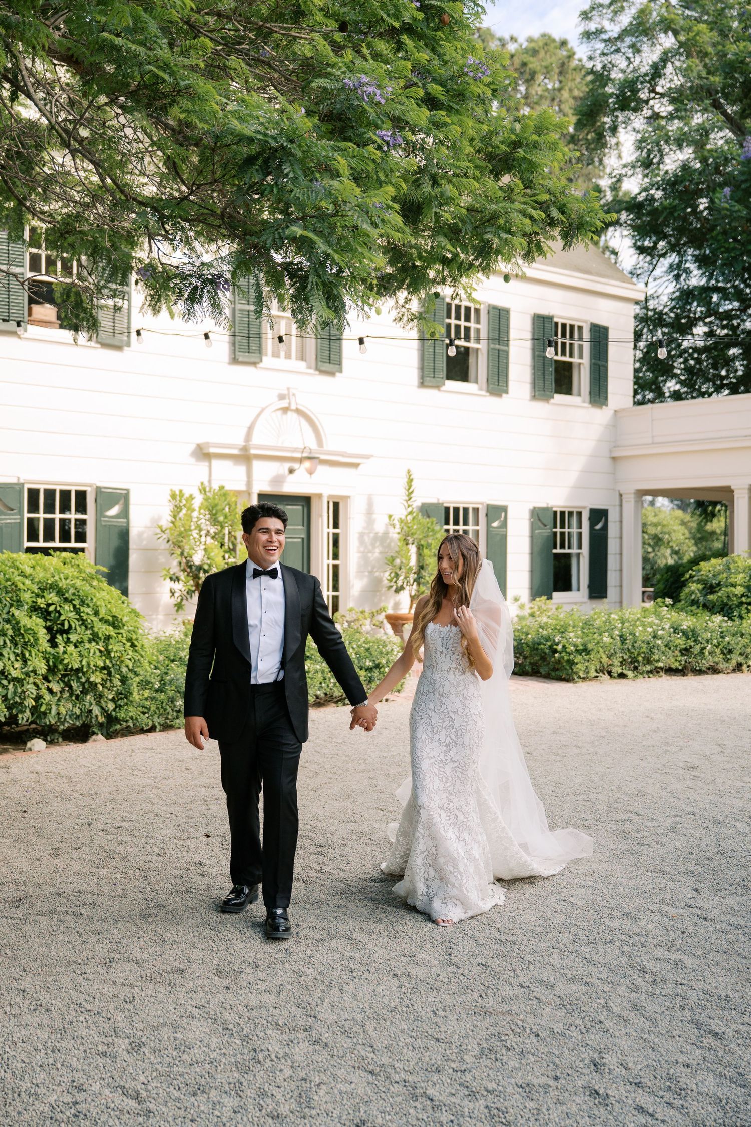 Bride and groom walk hand in hand outside elegant white manor with green shutters on their wedding day.