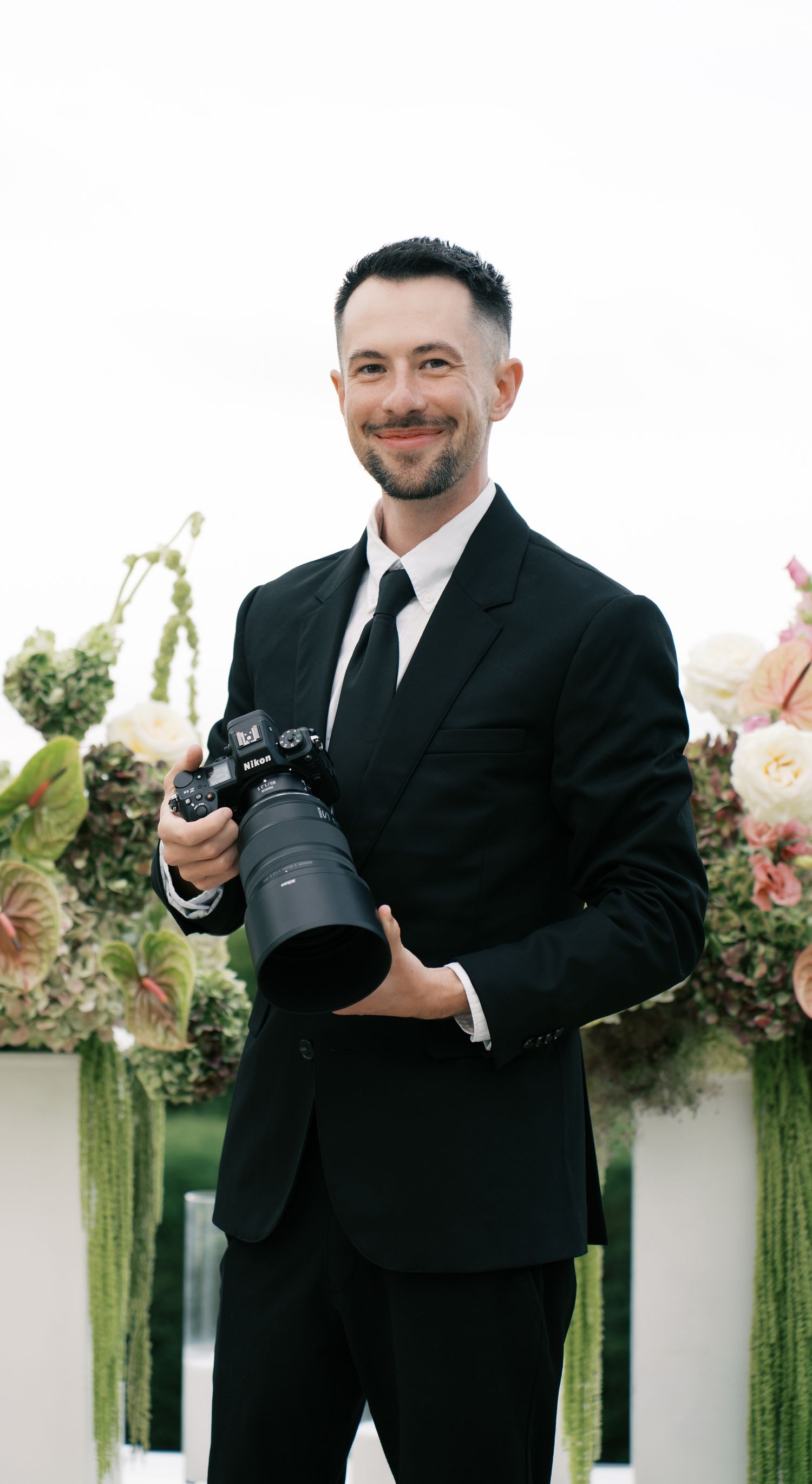 Professional photographer in a black suit holds a camera while smiling in a garden setting with white walls and greenery.