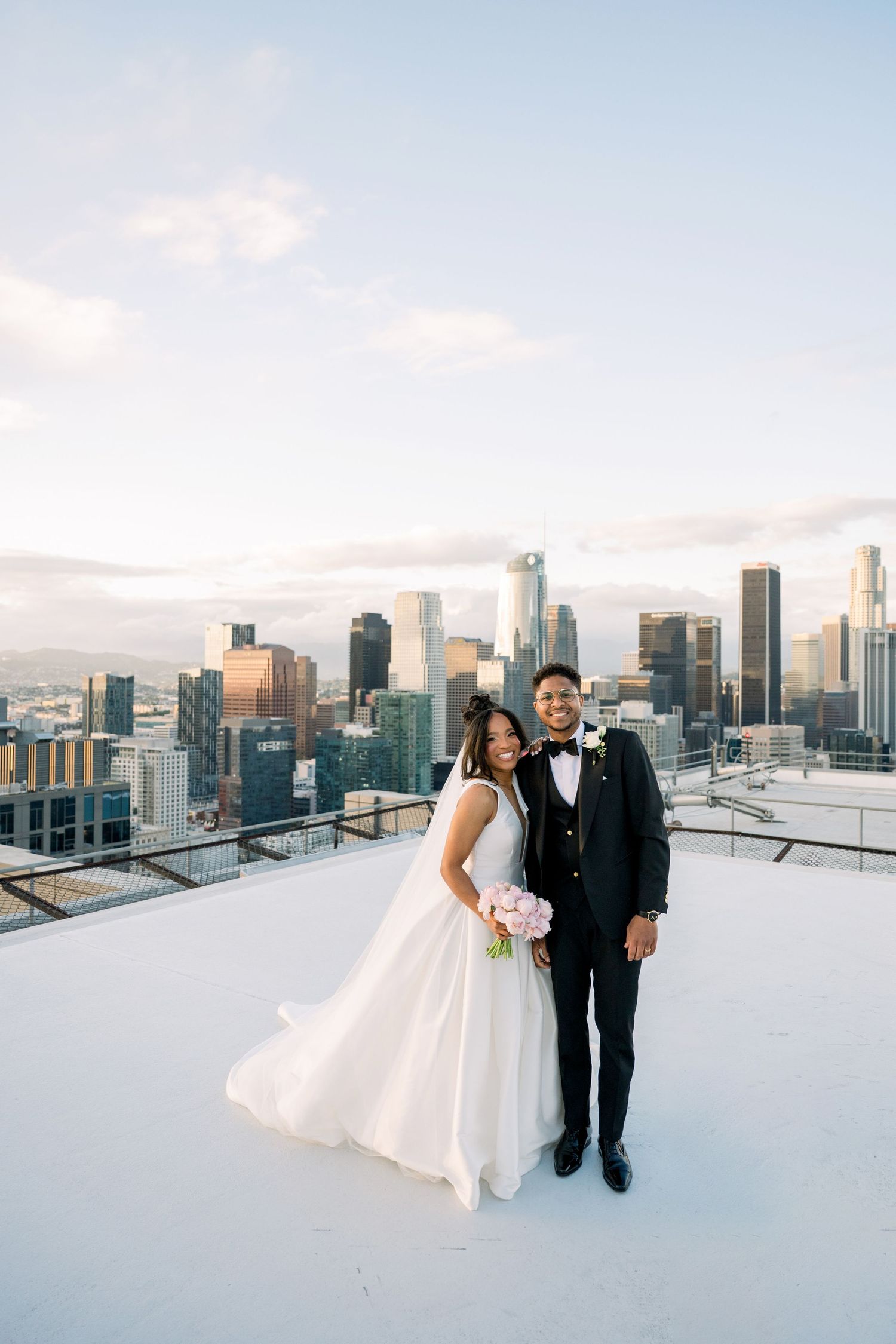 Newlywed couple stands on rooftop with downtown Los Angeles skyline at sunset in romantic wedding photo.