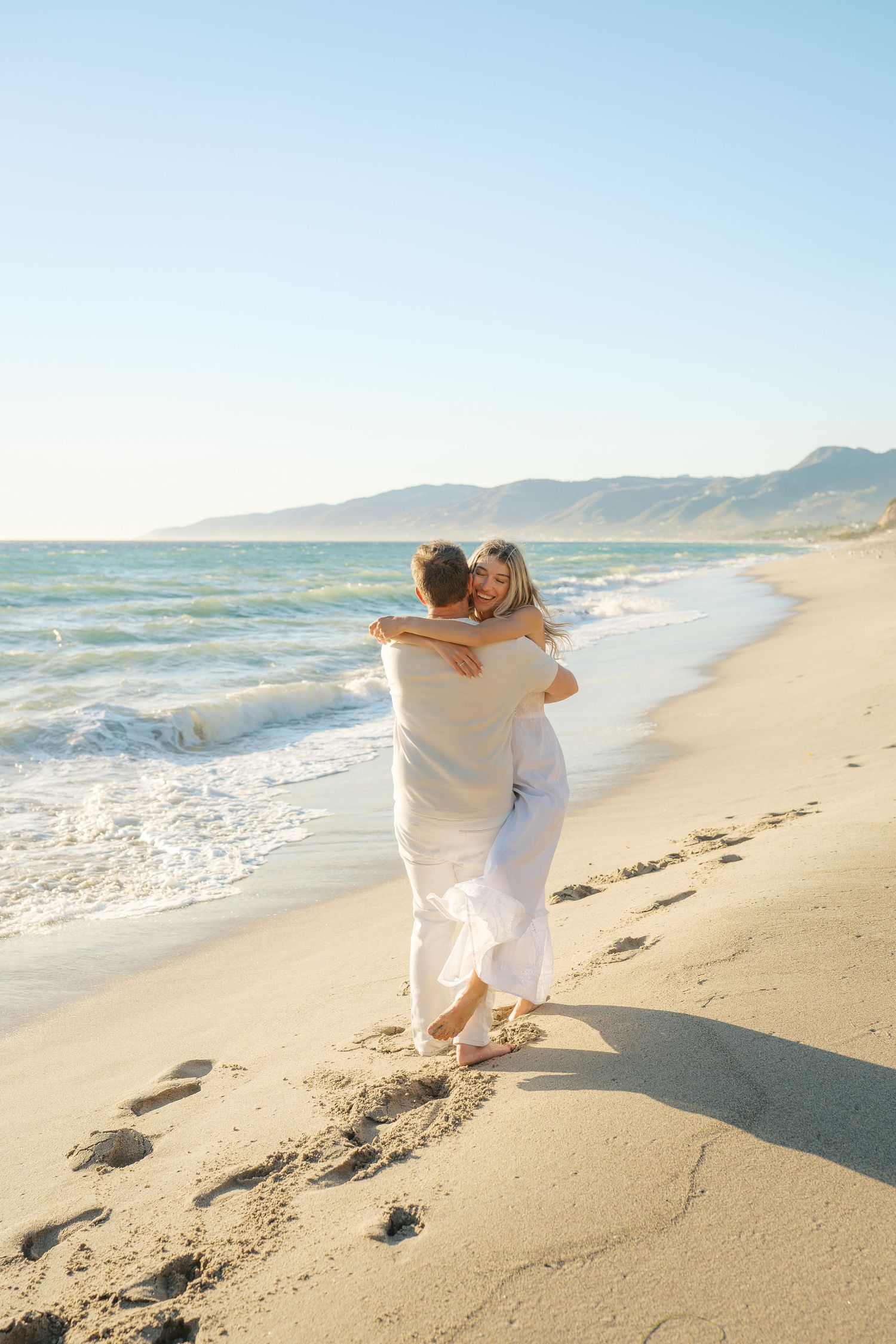 Couple embracing and walking barefoot on a sandy beach with ocean waves and mountains in the background.