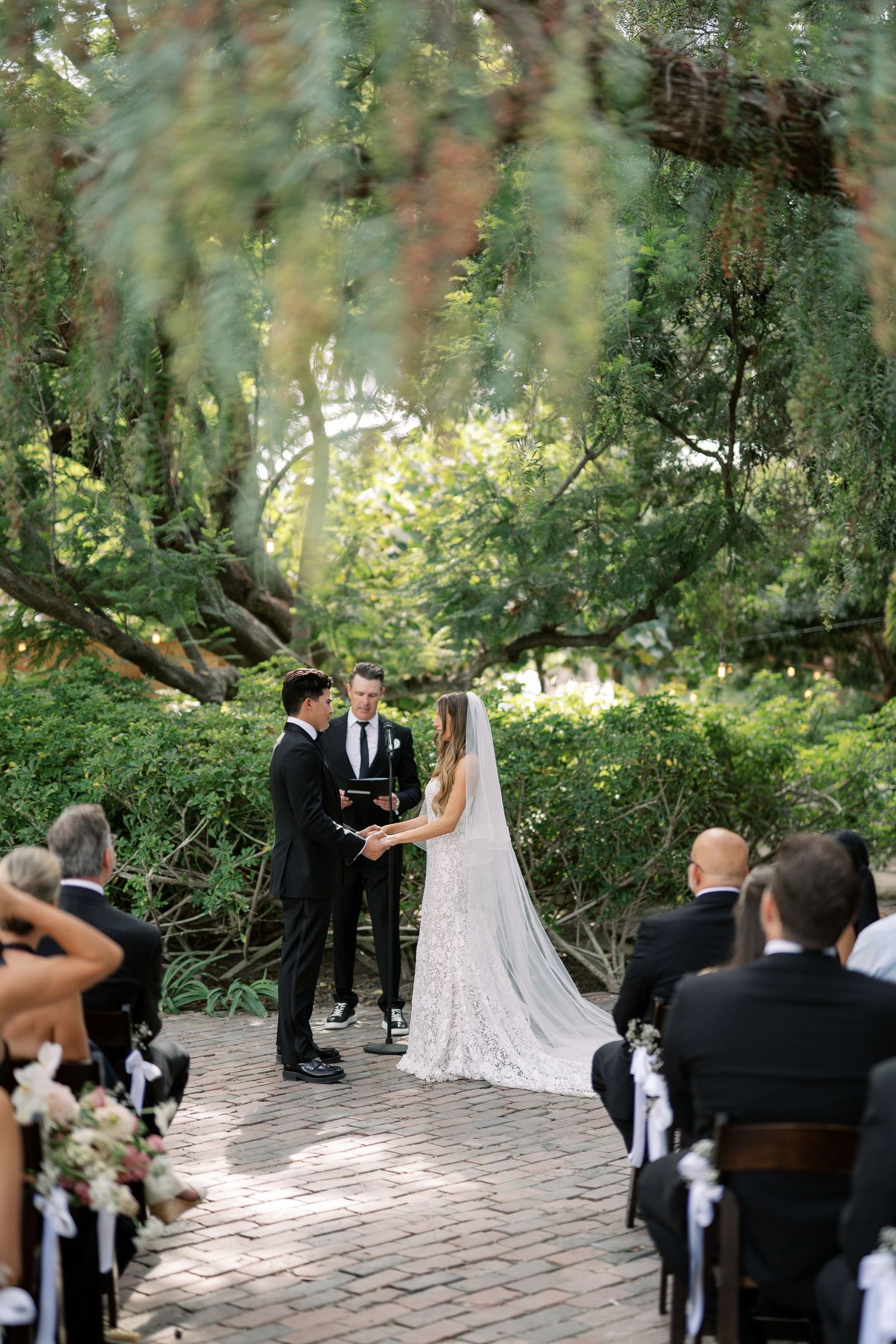 Outdoor wedding ceremony on brick path with bride and groom holding hands before officiant, surrounded by lush green trees.