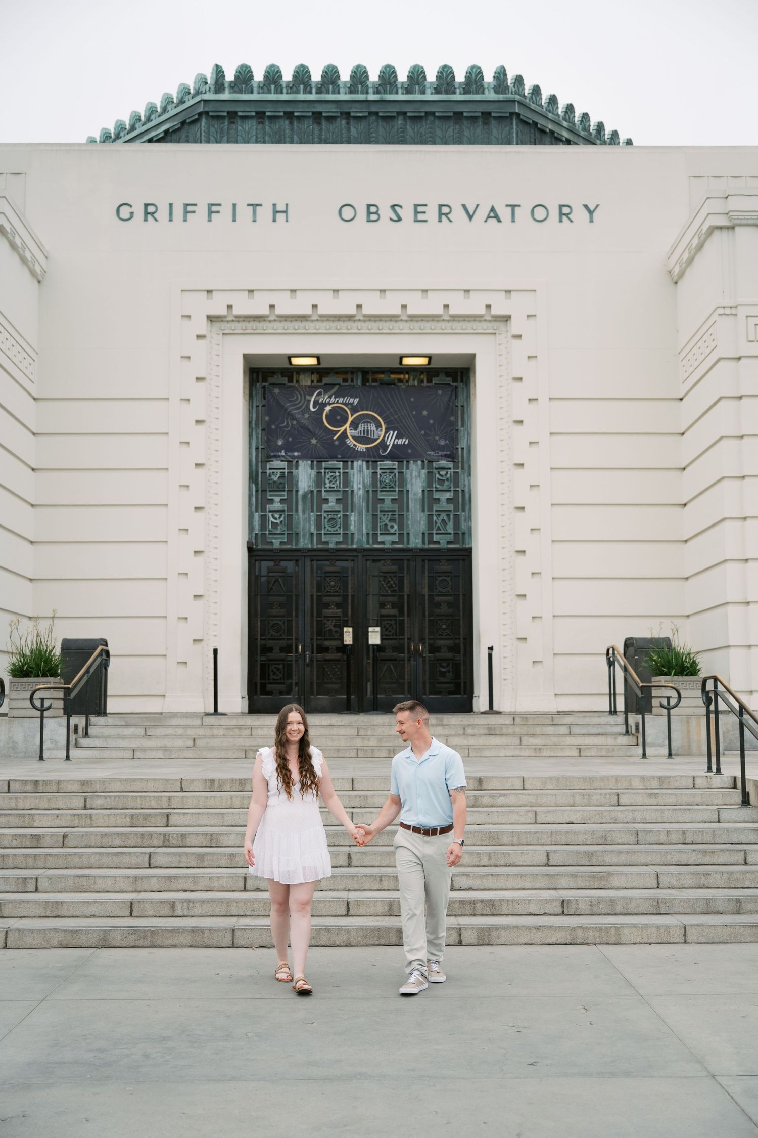 Two people stand together on the grand steps of the art deco Griffith Observatory building in Los Angeles.
