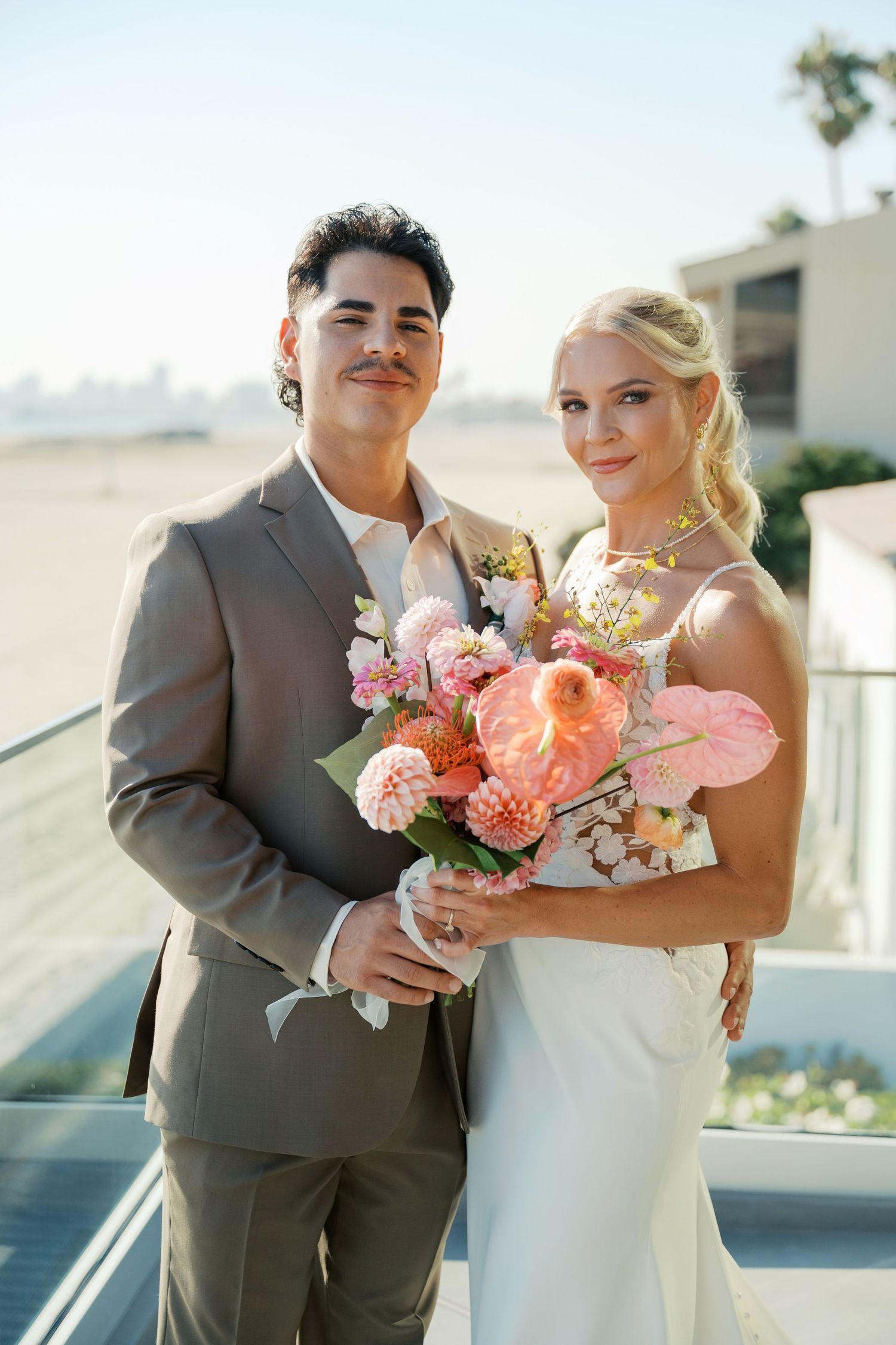 A couple in wedding attire poses together outdoors holding a coral and pink floral bouquet on a sunny day.