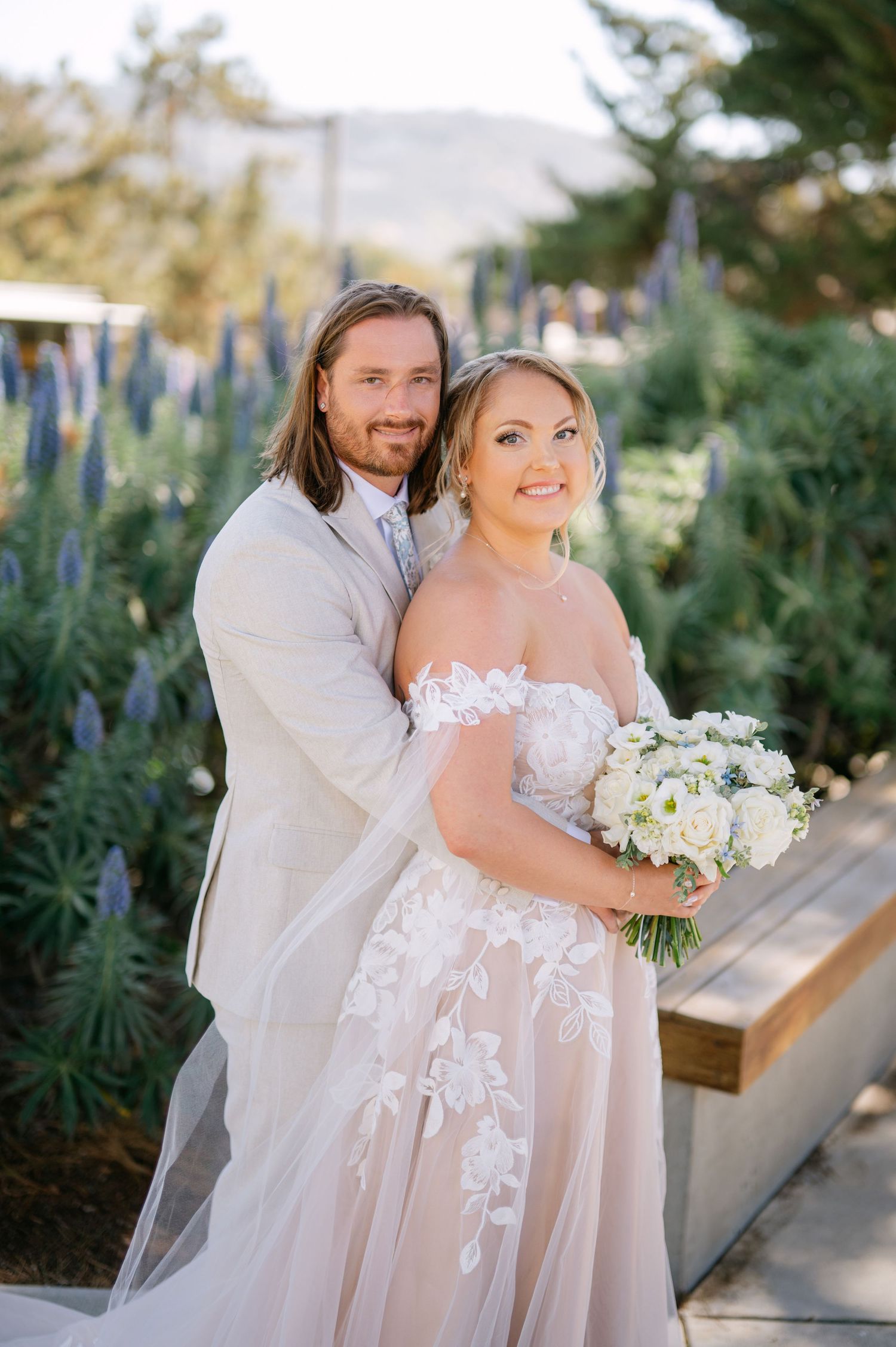 A couple posing outdoors on their wedding day in a garden setting with the bride holding a white floral bouquet.