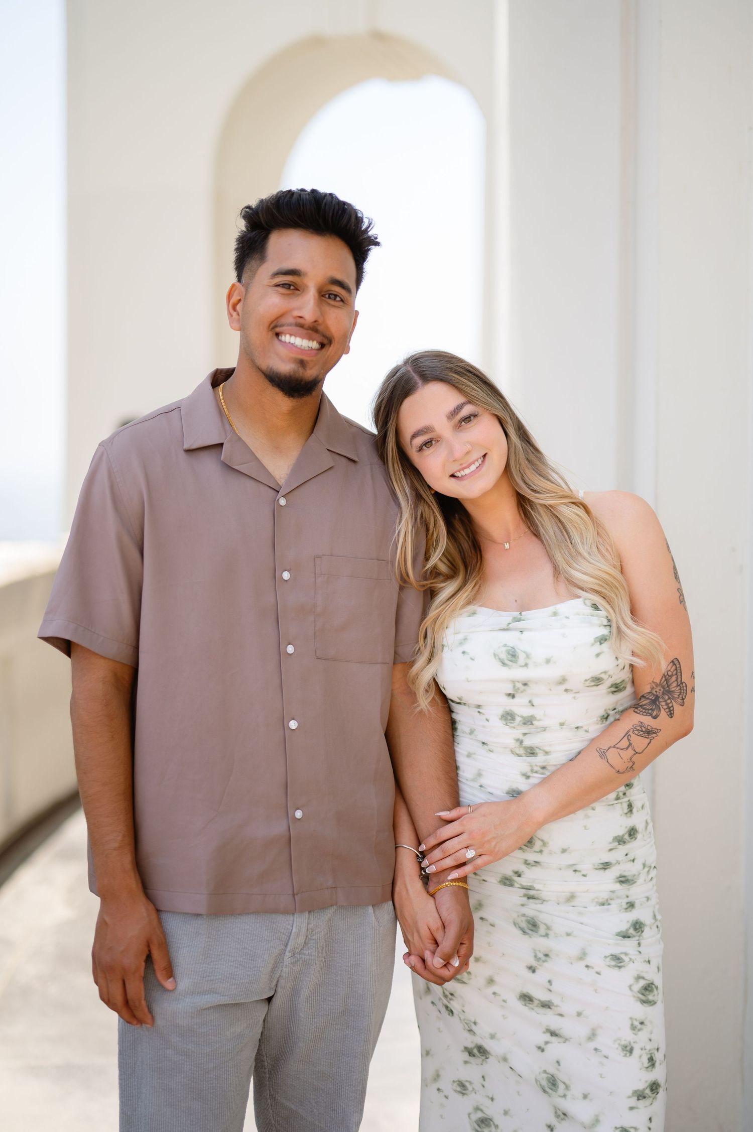 A couple poses together in a bright interior hallway wearing coordinated brown and white floral outfits.