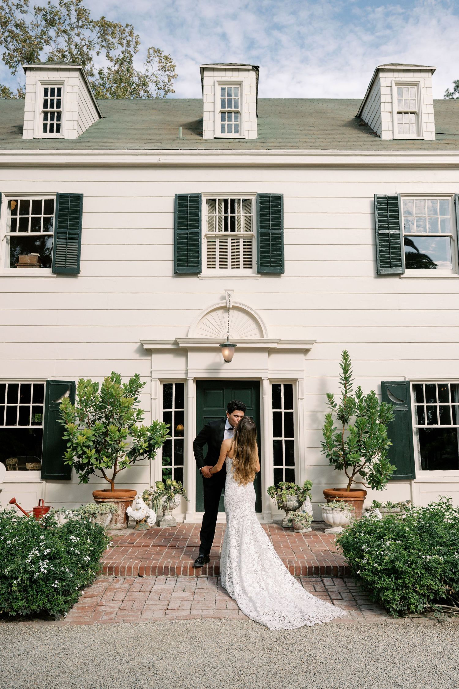 Bride and groom embracing in front of a white colonial mansion with green shutters and brick pathway on wedding day.