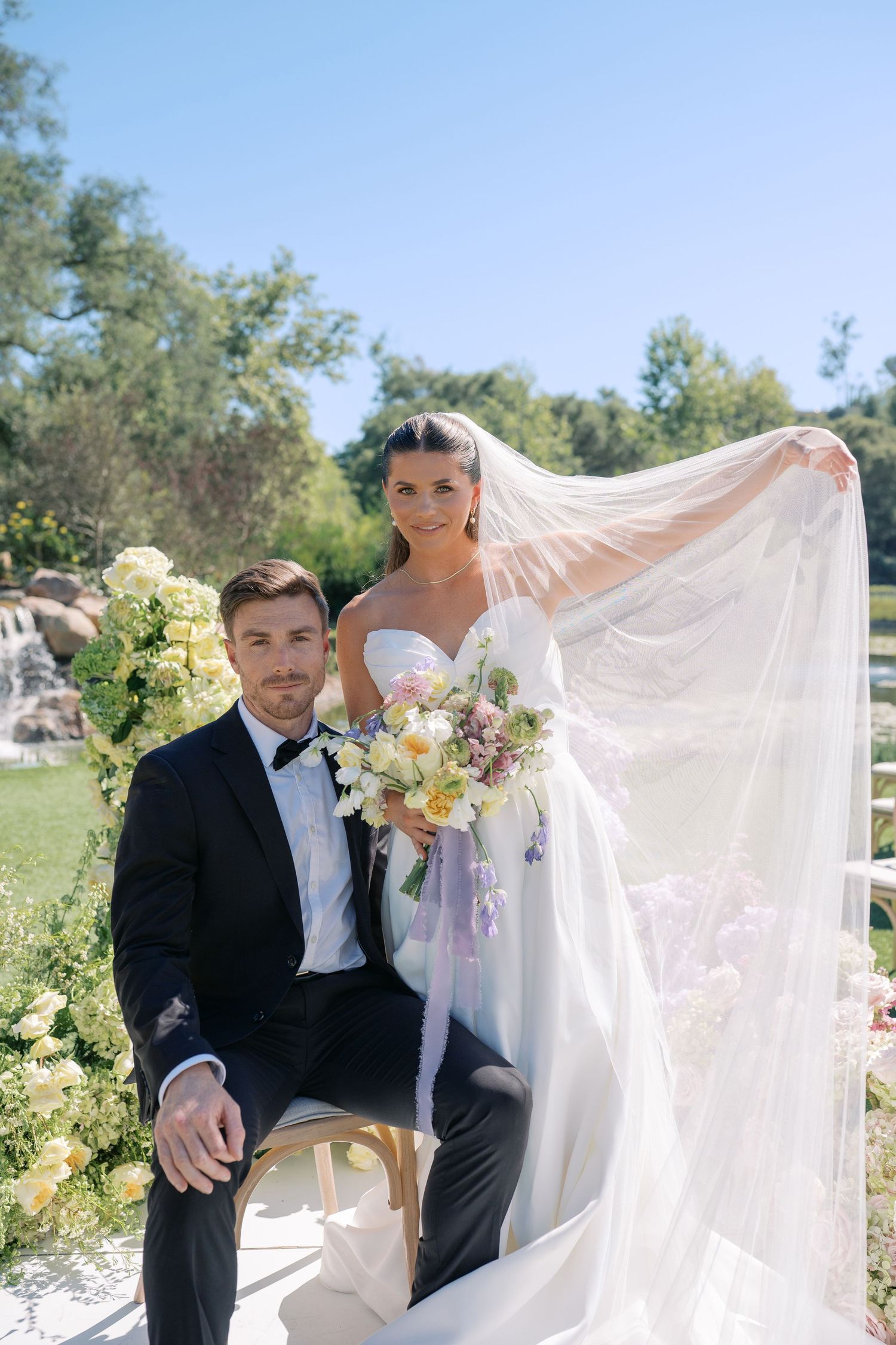 Elegant bride in strapless gown with flowing veil holds floral bouquet beside groom in black tuxedo at outdoor wedding.