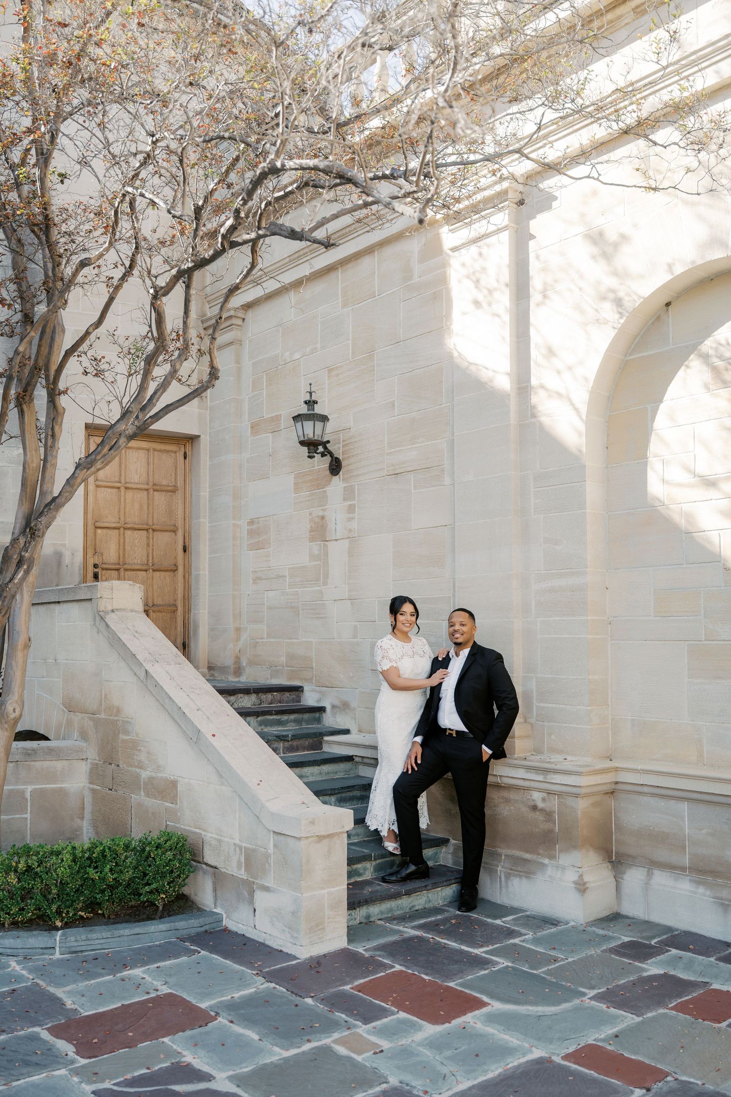 A couple poses in an elegant stone archway courtyard with stone steps and greenery in the background.