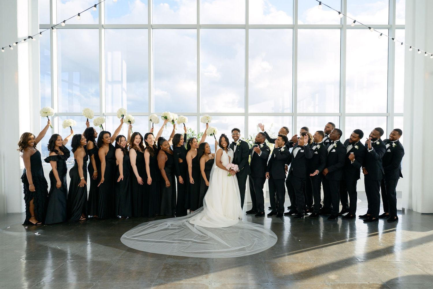 A large wedding party in formal attire poses together against floor-to-ceiling windows.