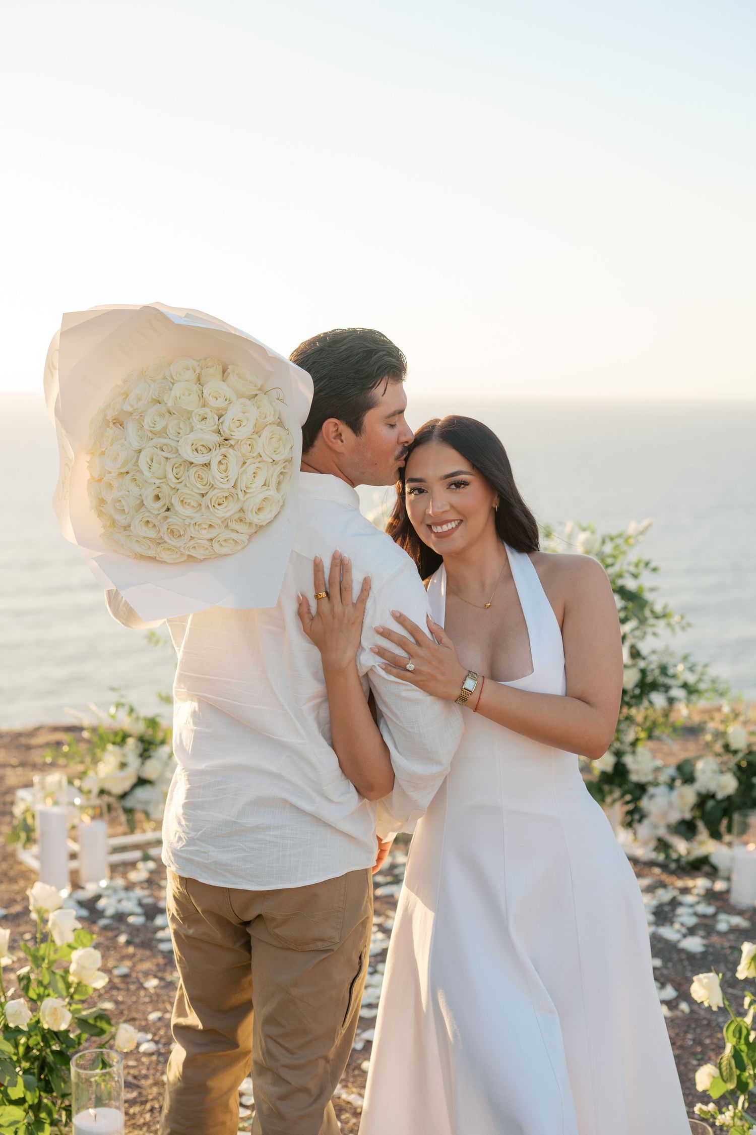 Couple celebrating engagement at sunset cliffside, woman in white dress showing ring, holding large white rose bouquet by the ocean.