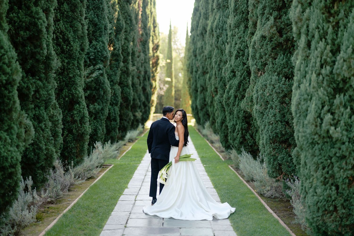 Bride and groom share a romantic kiss along an elegant tree-lined garden path on their wedding day.
