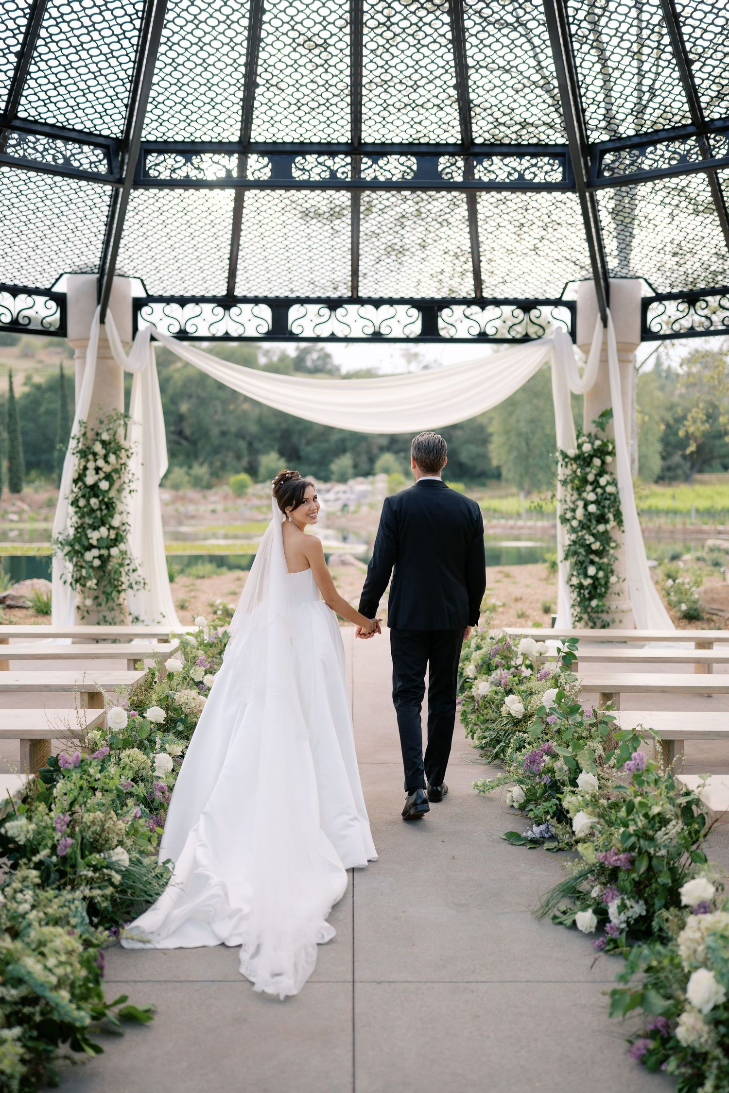 Bride and groom walk hand-in-hand under an elegant gazebo adorned with white draping and floral arrangements at their wedding.