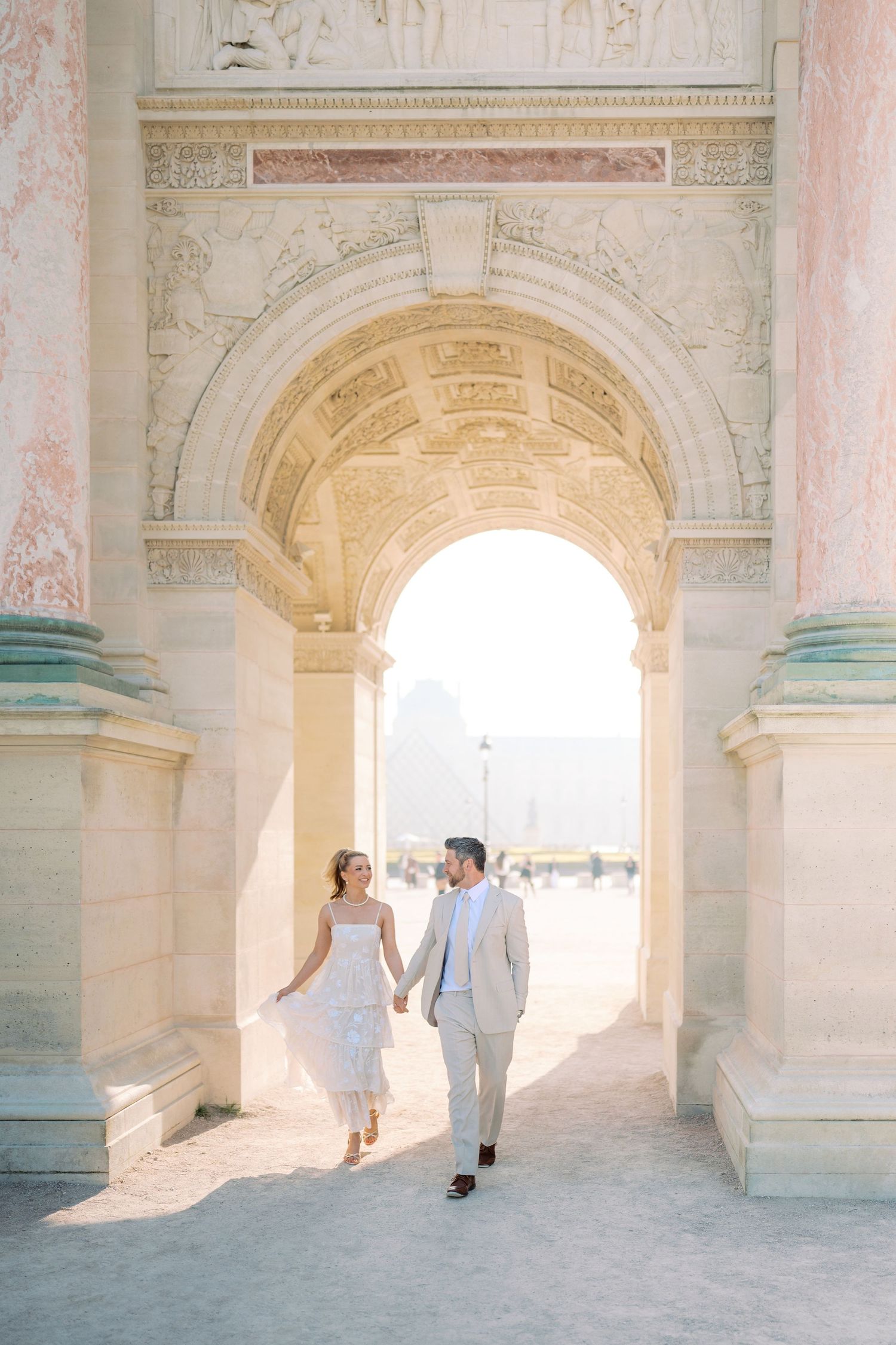 Couple holding hands walking through ornate arch at the Louvre in Paris, bride in white dress, groom in tan suit.