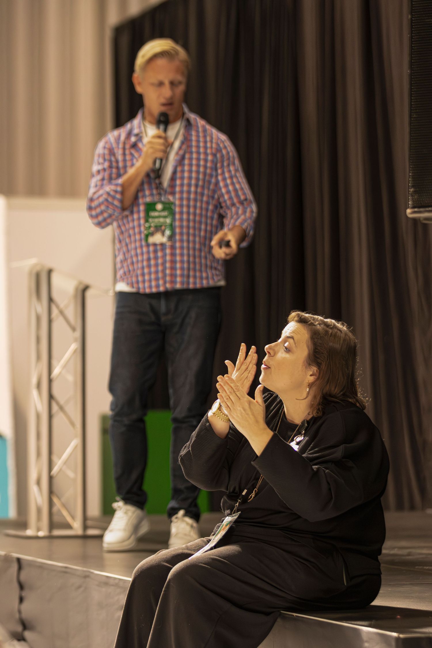 Person in a black hoodie performs on stage while someone in a plaid shirt stands in the background.