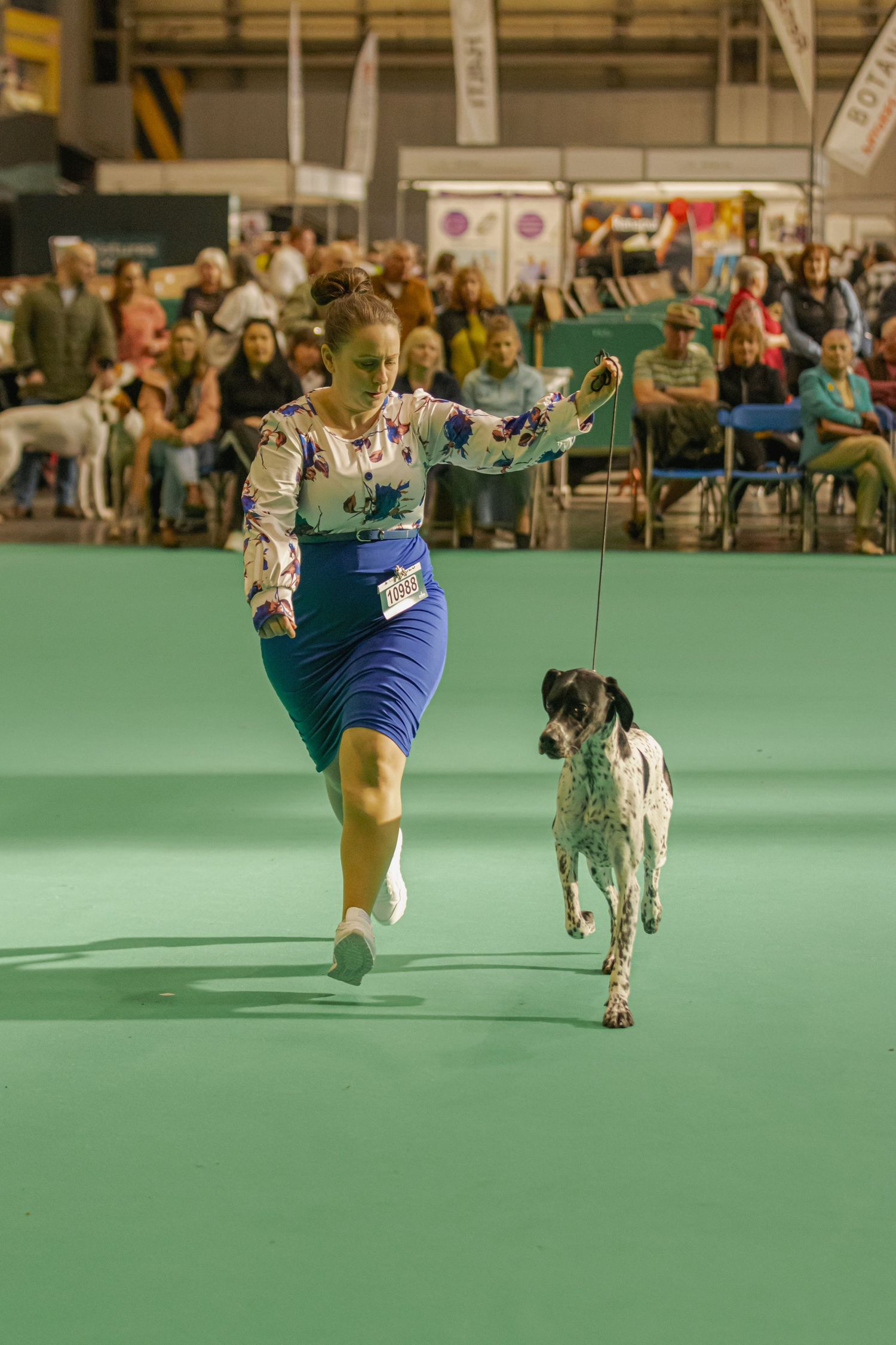 Dog and handler perform synchronized dance routine on green carpet at indoor dog show competition.