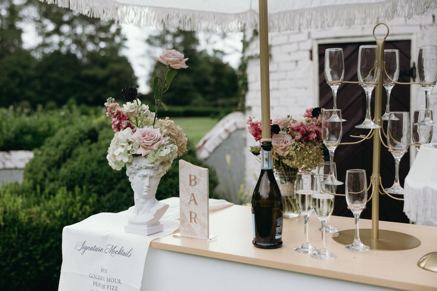 Elegant outdoor wedding bar setup with champagne flutes, floral arrangements, and a BAR sign on a white draped table.