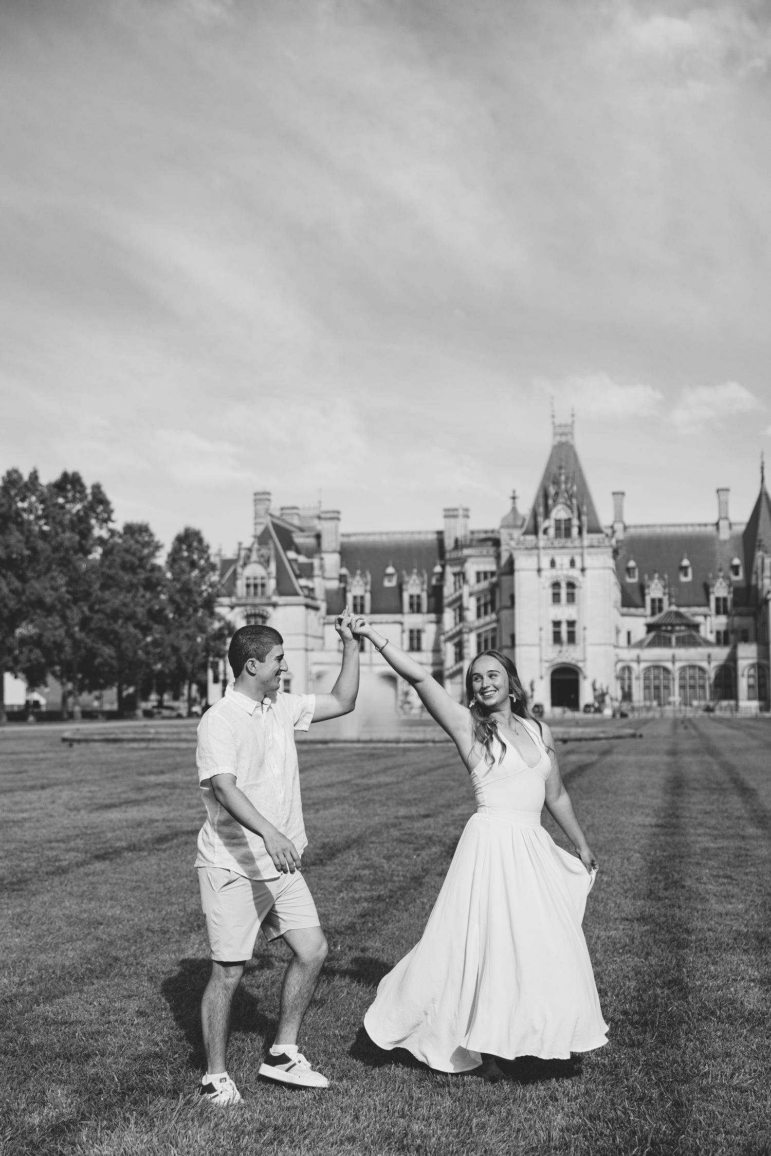 Black and white photo of a couple dancing on a lawn in front of a grand estate building under a cloudy sky.