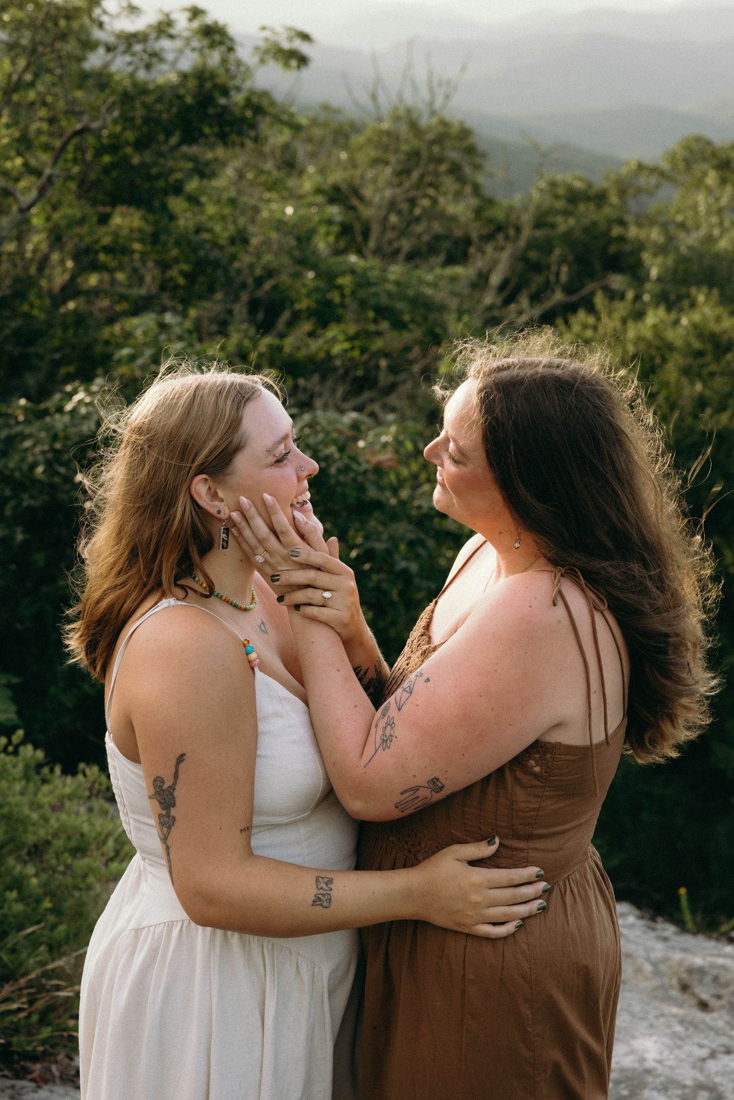 Two women with tattoos embrace tenderly outdoors, one in white and one in brown dress, with lush green mountains behind them.