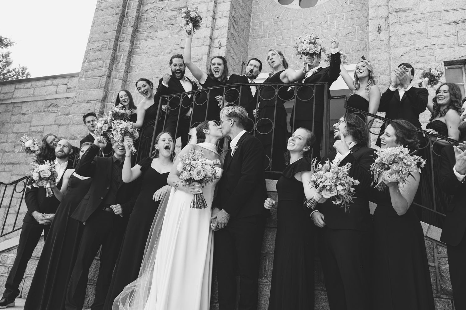 Black and white wedding party photo on church steps with bride and groom kissing surrounded by cheering bridesmaids and groomsmen.