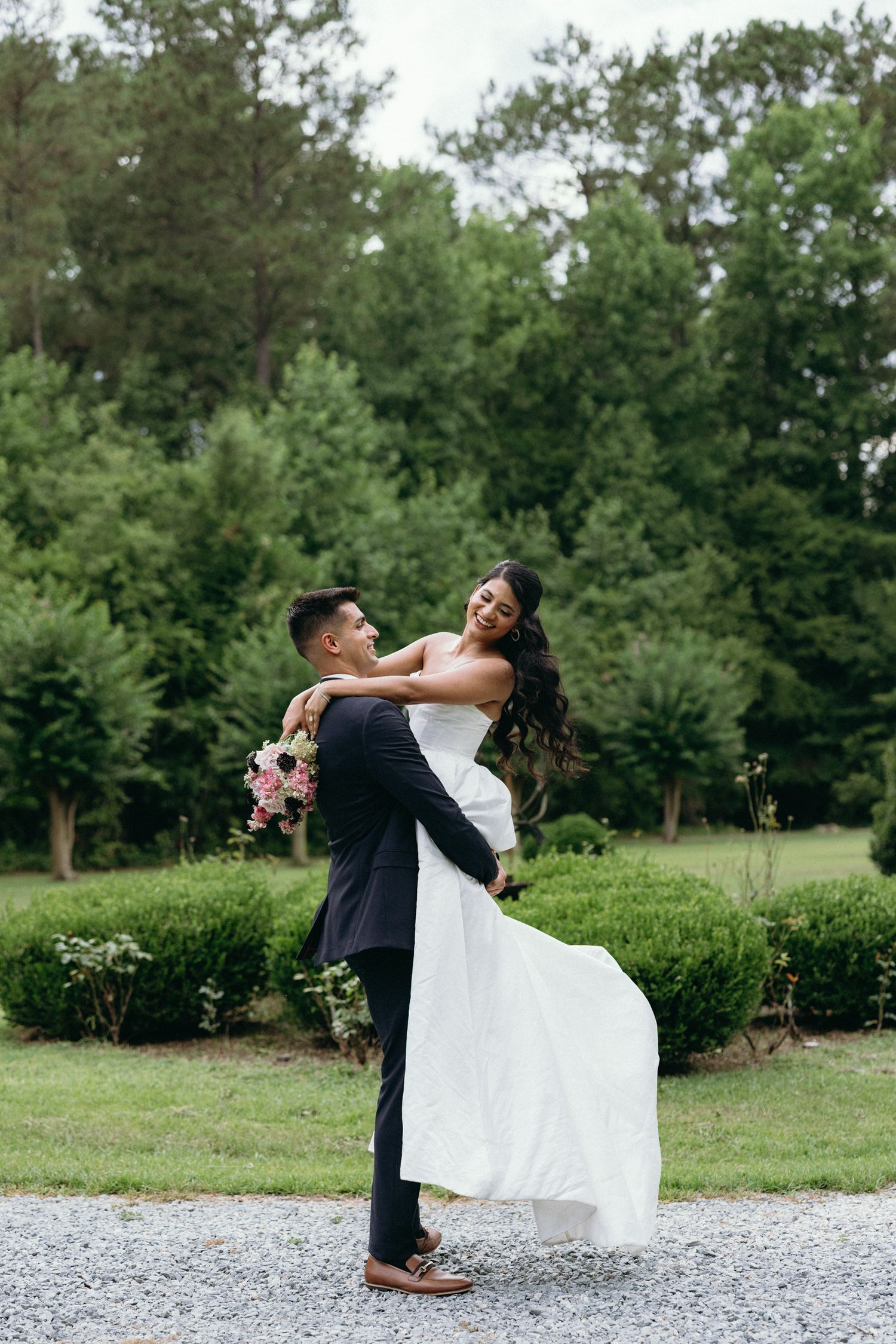 Groom lifting smiling bride in white gown outdoors, surrounded by lush green trees and manicured shrubs on wedding day.
