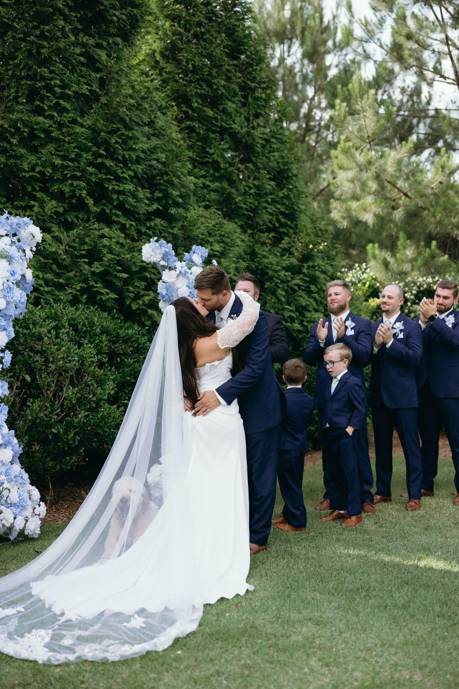 Wedding party in navy suits and white dress gathered outdoors among greenery. The Bradford, Separk  Mansion, Graylyn Estate