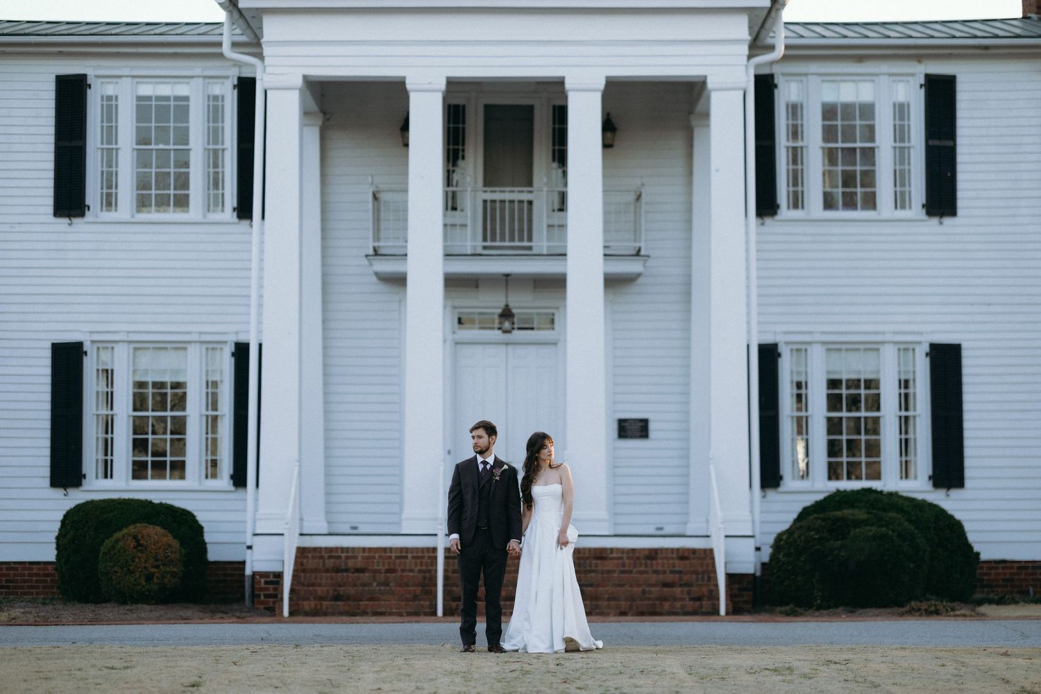 Bride and groom stand before a grand white colonial mansion with black shutters and columns on their wedding day.