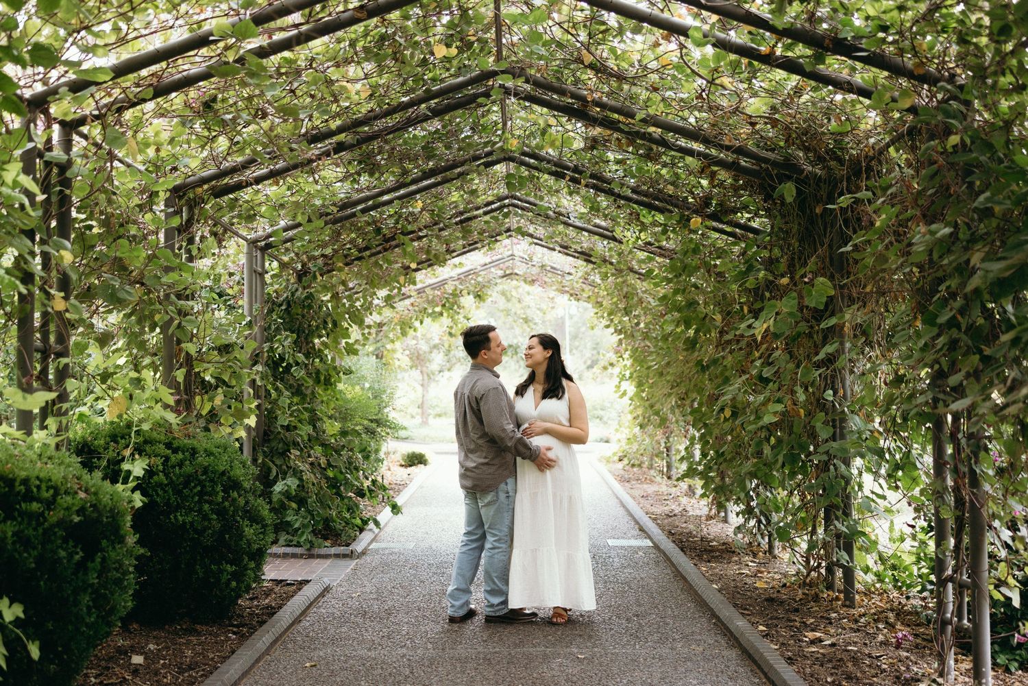 Couple stands facing each other under a lush green garden arbor tunnel on a stone pathway.