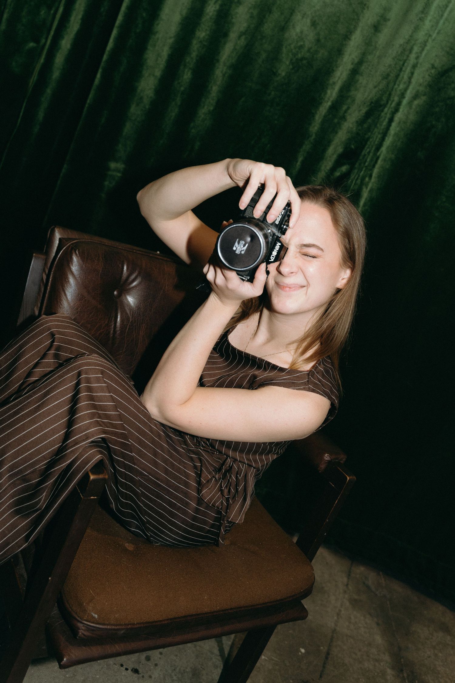 A photographer in a striped dress reclines on a brown leather chair while taking a photo with a professional camera.
