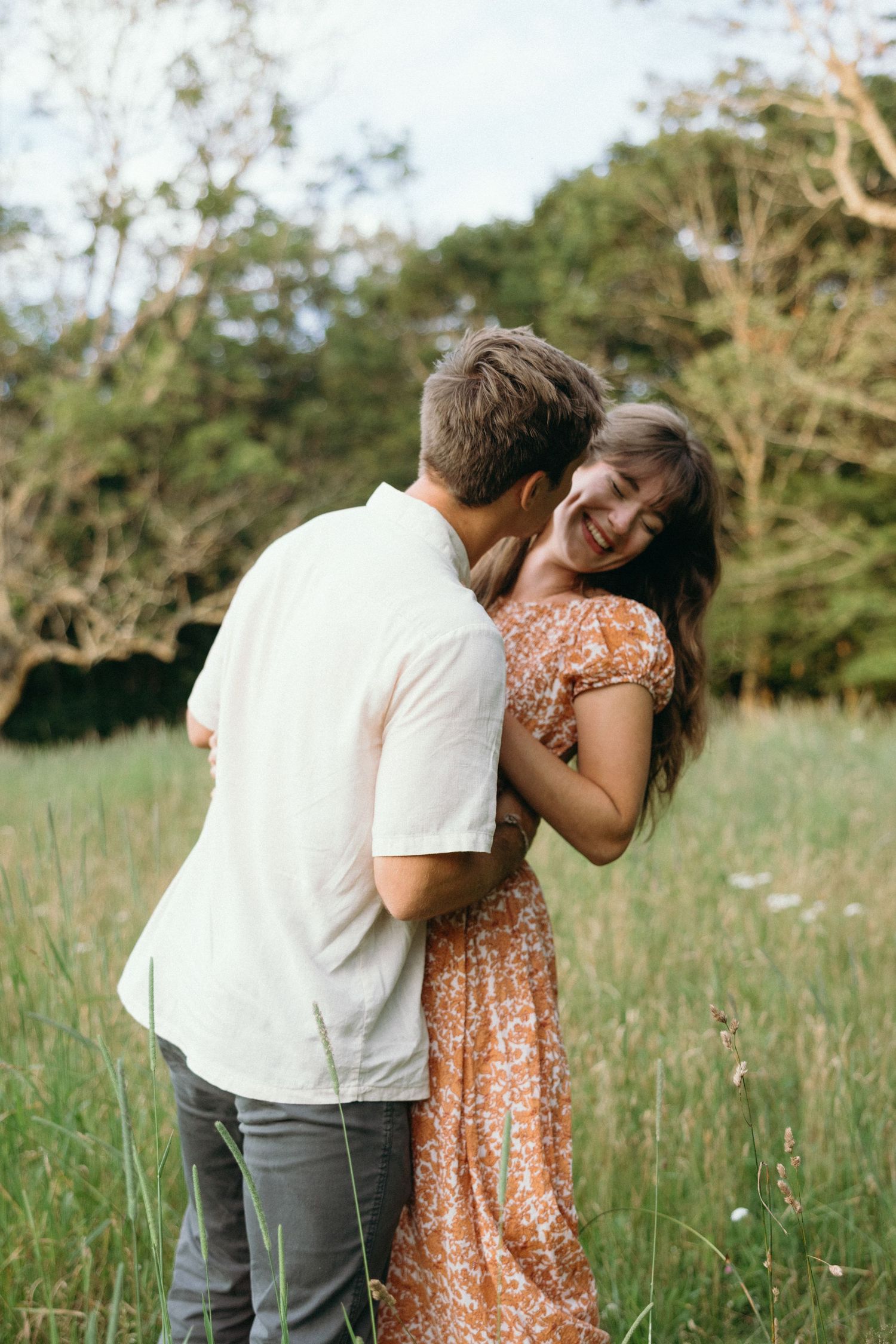 A couple shares an intimate moment in a meadow, with her wearing a floral peach dress and him in a white shirt.