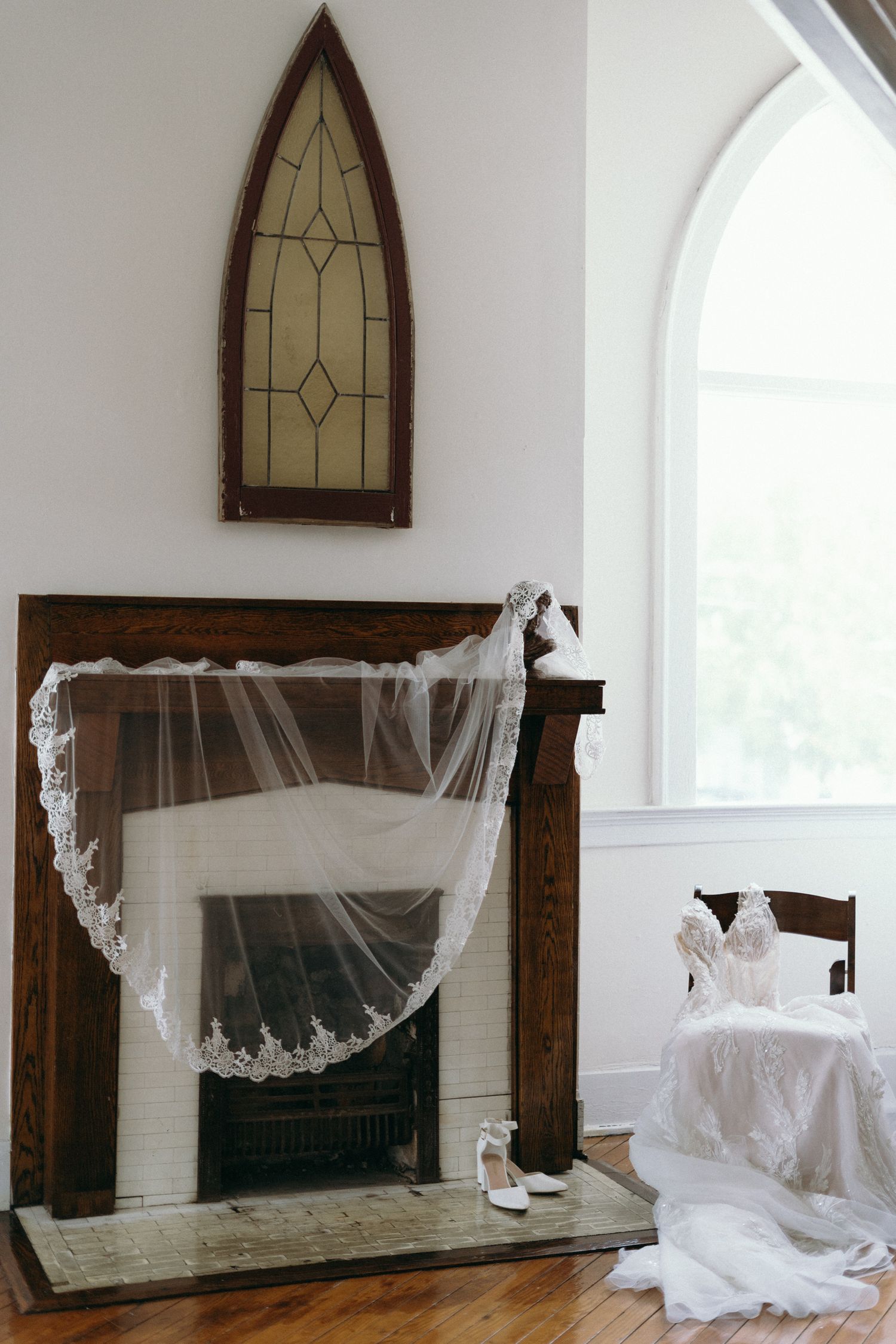 Bridal veil draped over rustic fireplace with wedding dress on chair and heels on floor in elegant chapel setting.