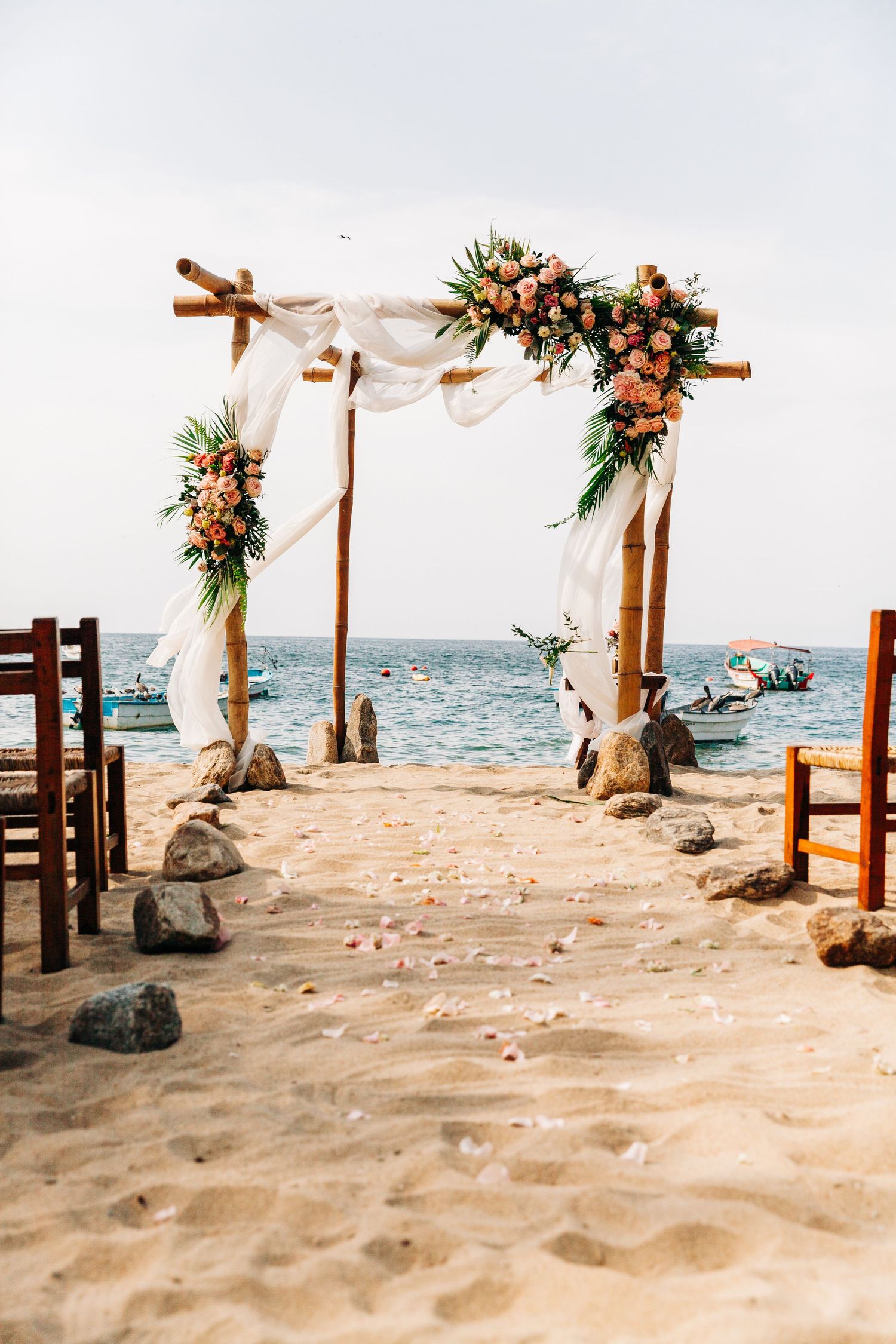 A rustic wooden wedding arch decorated with peach flowers and white fabric stands on a sandy beach overlooking the ocean.