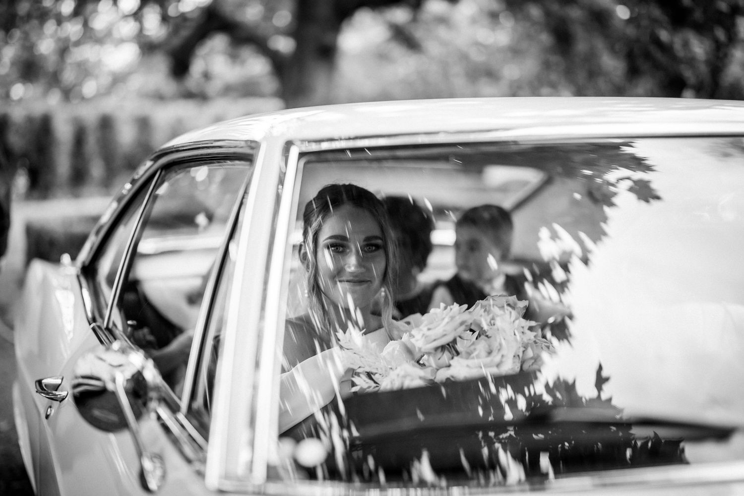 Monochrome portrait through car window showing wedding day transportation moment.