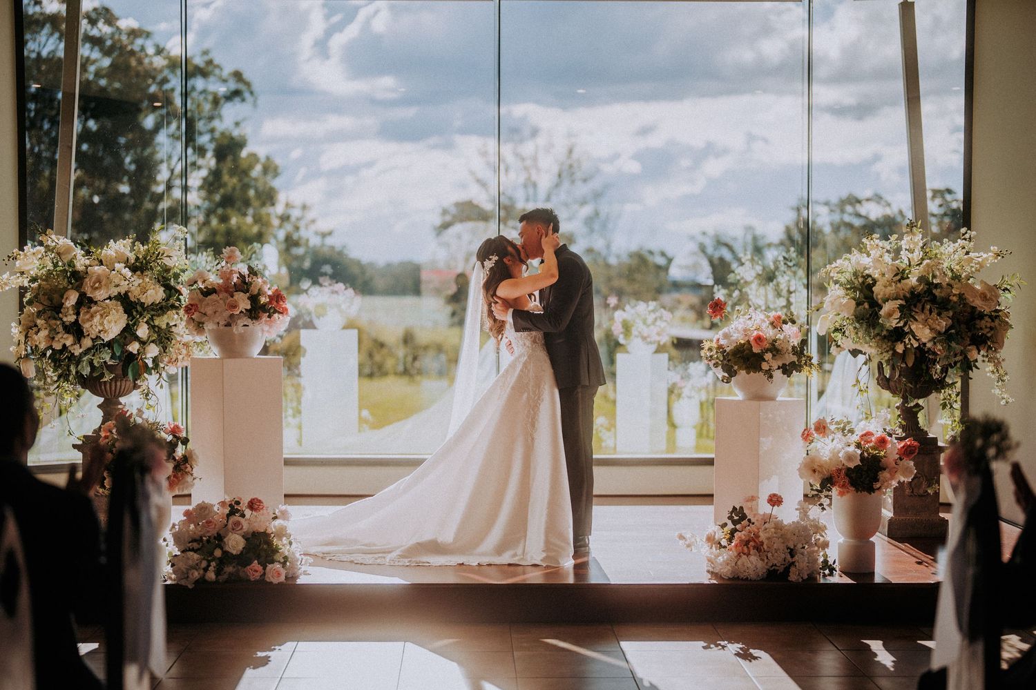 Wedding ceremony in elegant venue with floral arrangements framing couple by large windows.