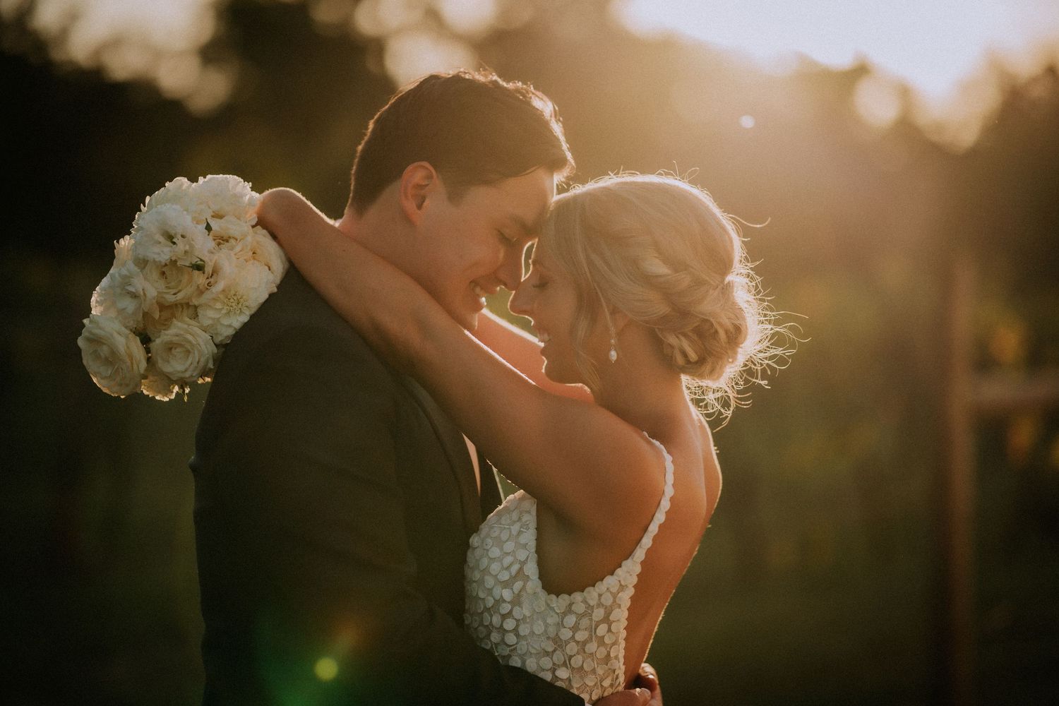 Romantic sunset wedding portrait with bride holding white bouquet during intimate embrace.