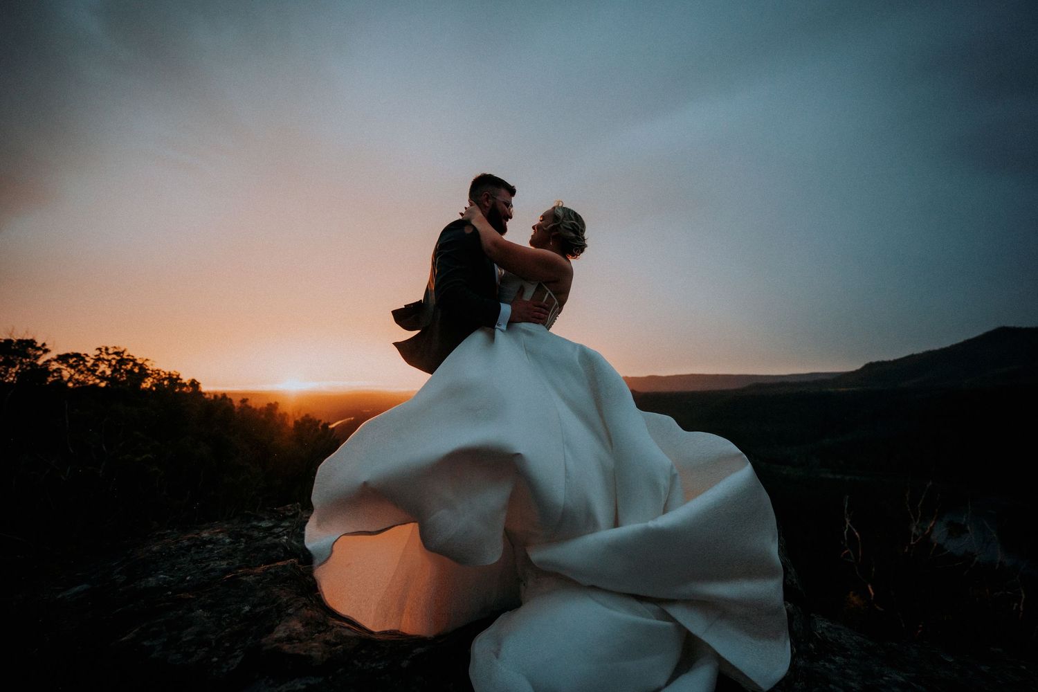 Newlyweds embrace against a sunset mountain backdrop as the bride's white dress flows in the wind.